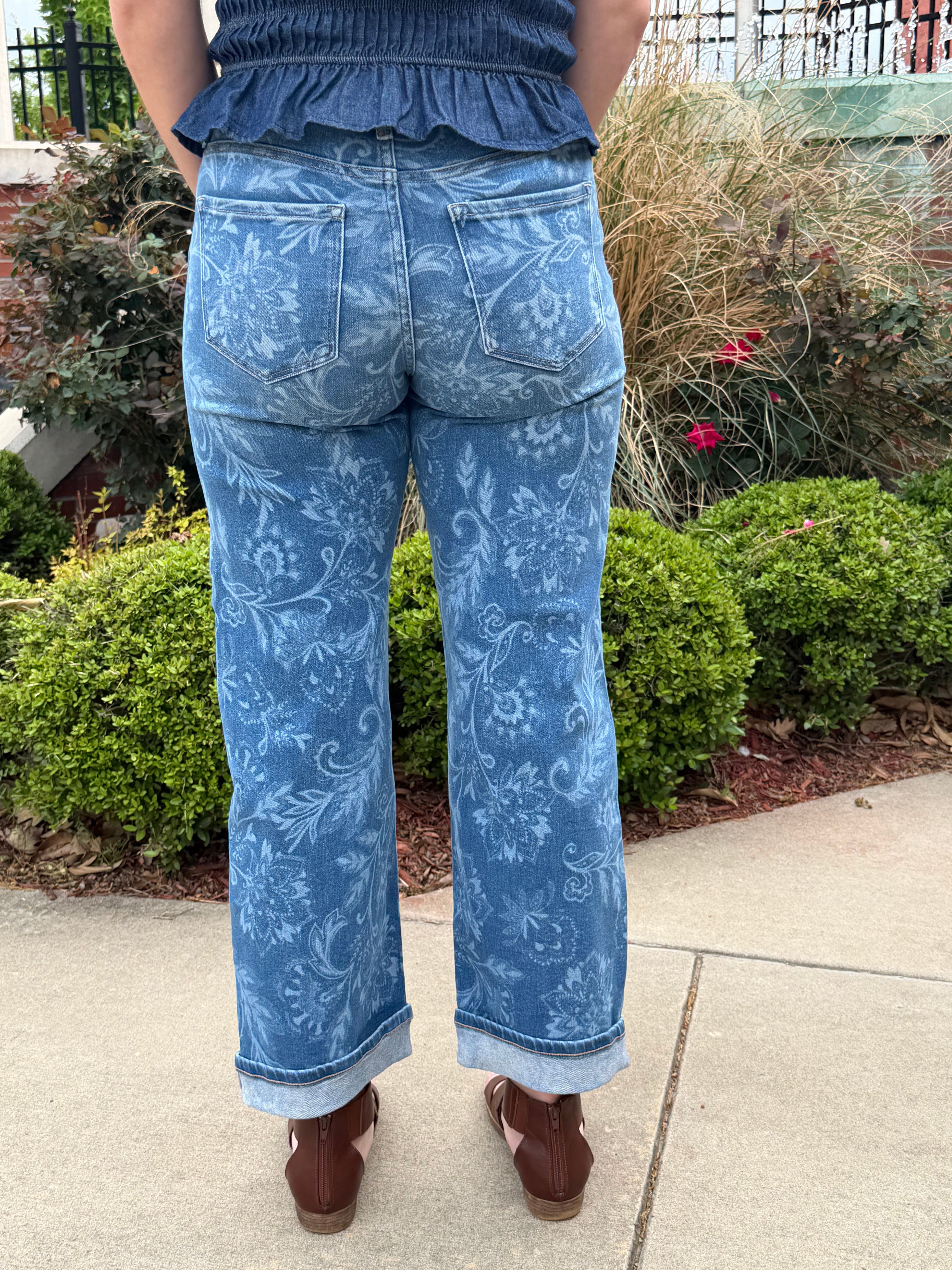 Person wearing blue floral-patterned jeans standing outdoors on a sidewalk.