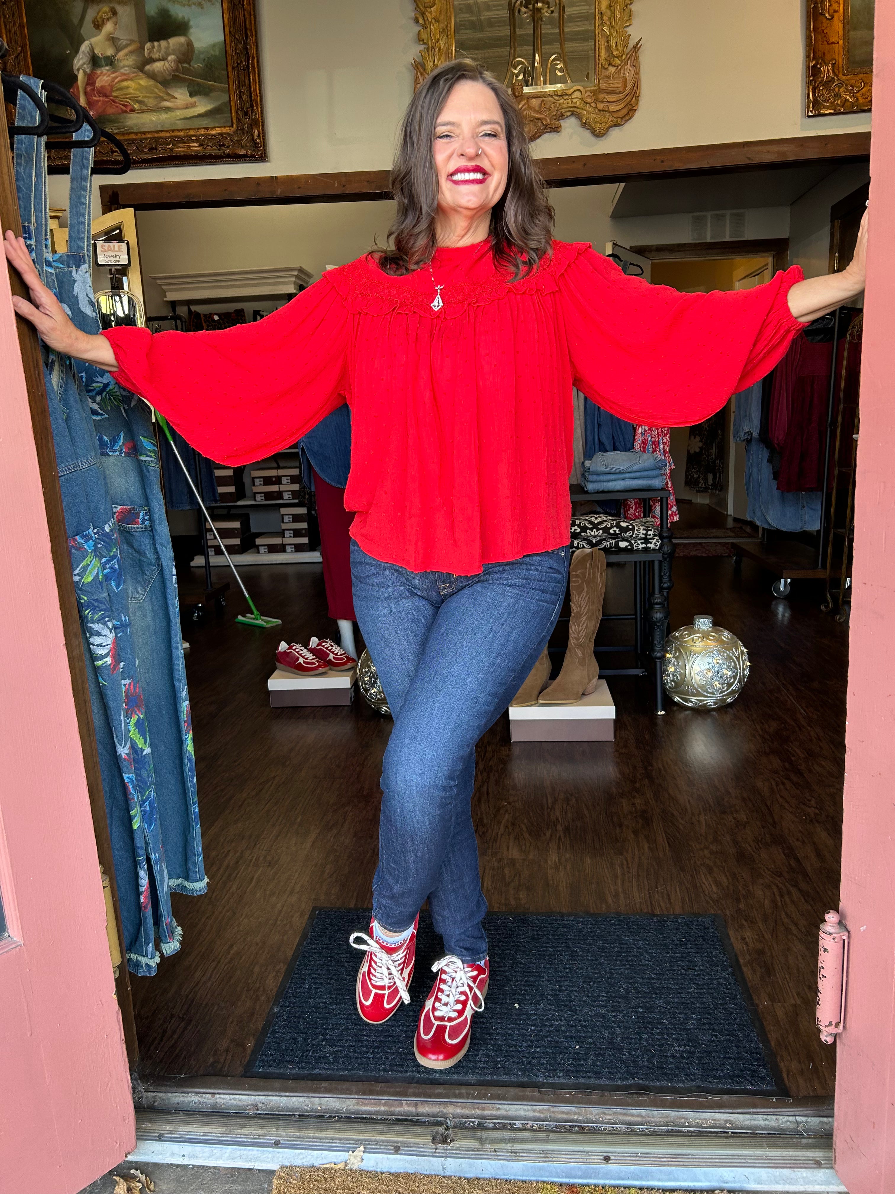 Woman wearing a red blouse and blue jeans standing in a doorway with a store interior in the background.