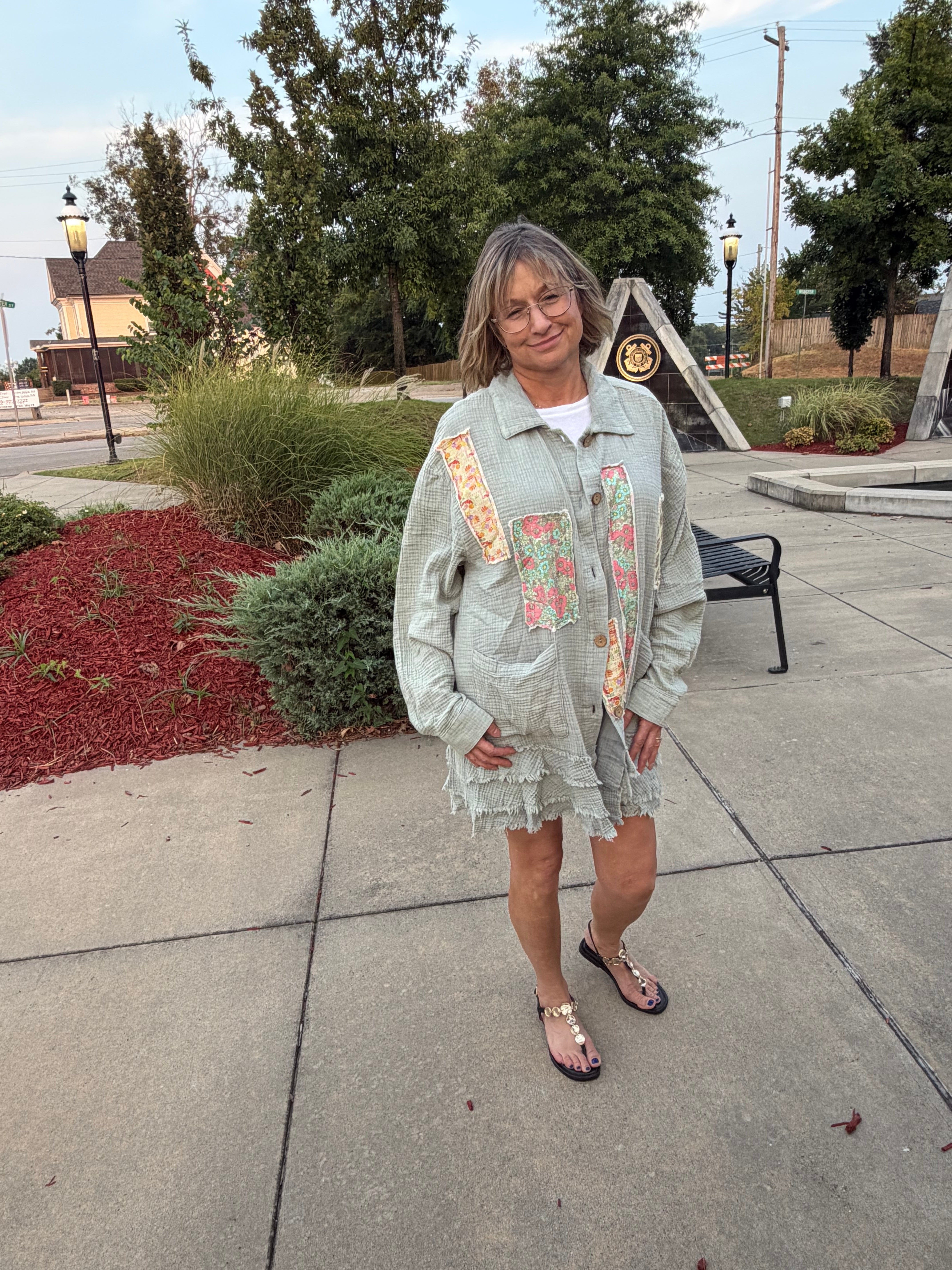 Front three-quarter view of woman wearing Blue Velvet sage patchwork gauze shirt showing floral patchwork front panels and tiered ruffle hem, outdoor park setting