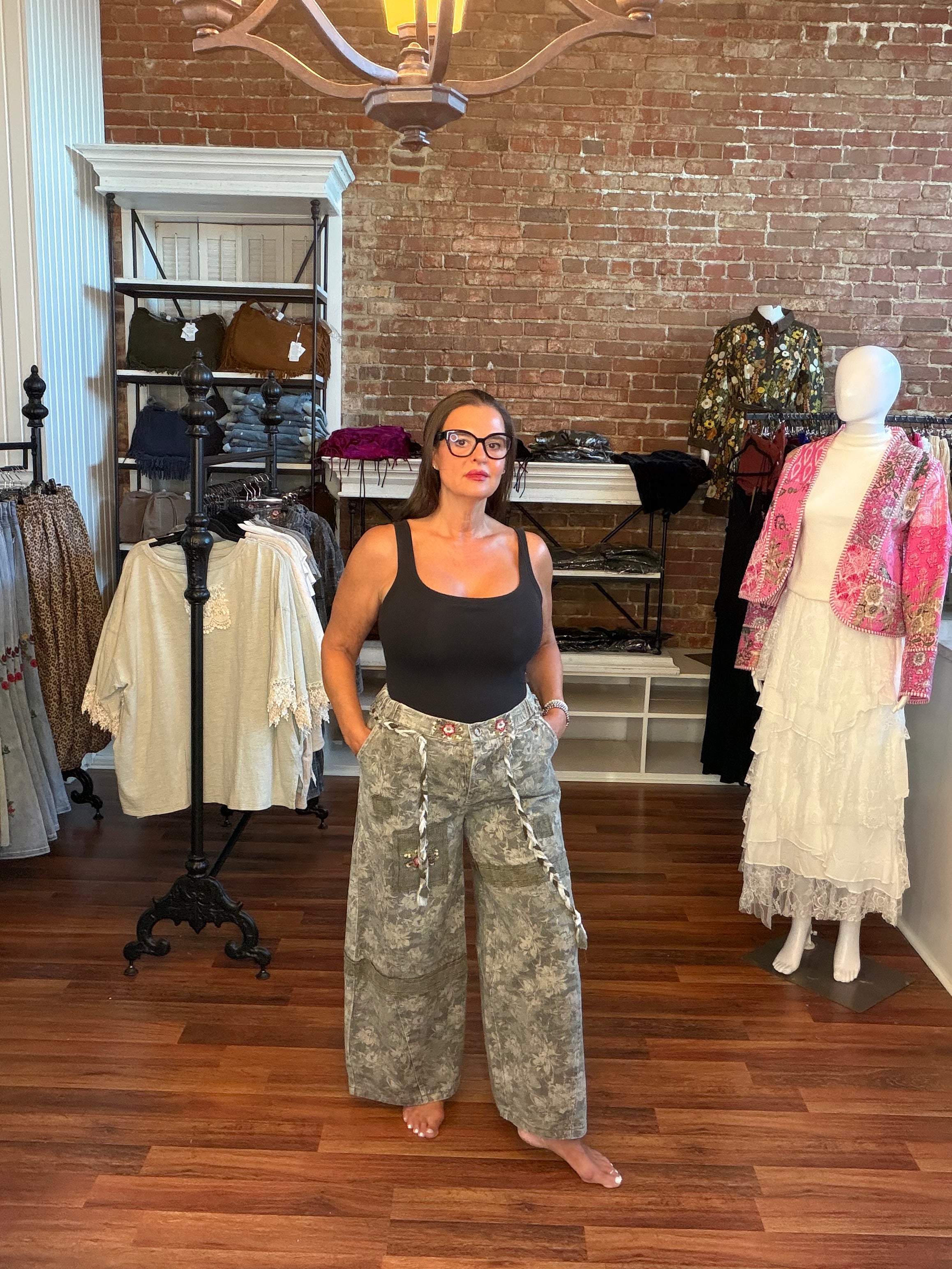 A woman wearing a black tank top and camouflage cargo pants stands barefoot in a boutique with a brick wall and wood floor, surrounded by clothing racks and mannequins.