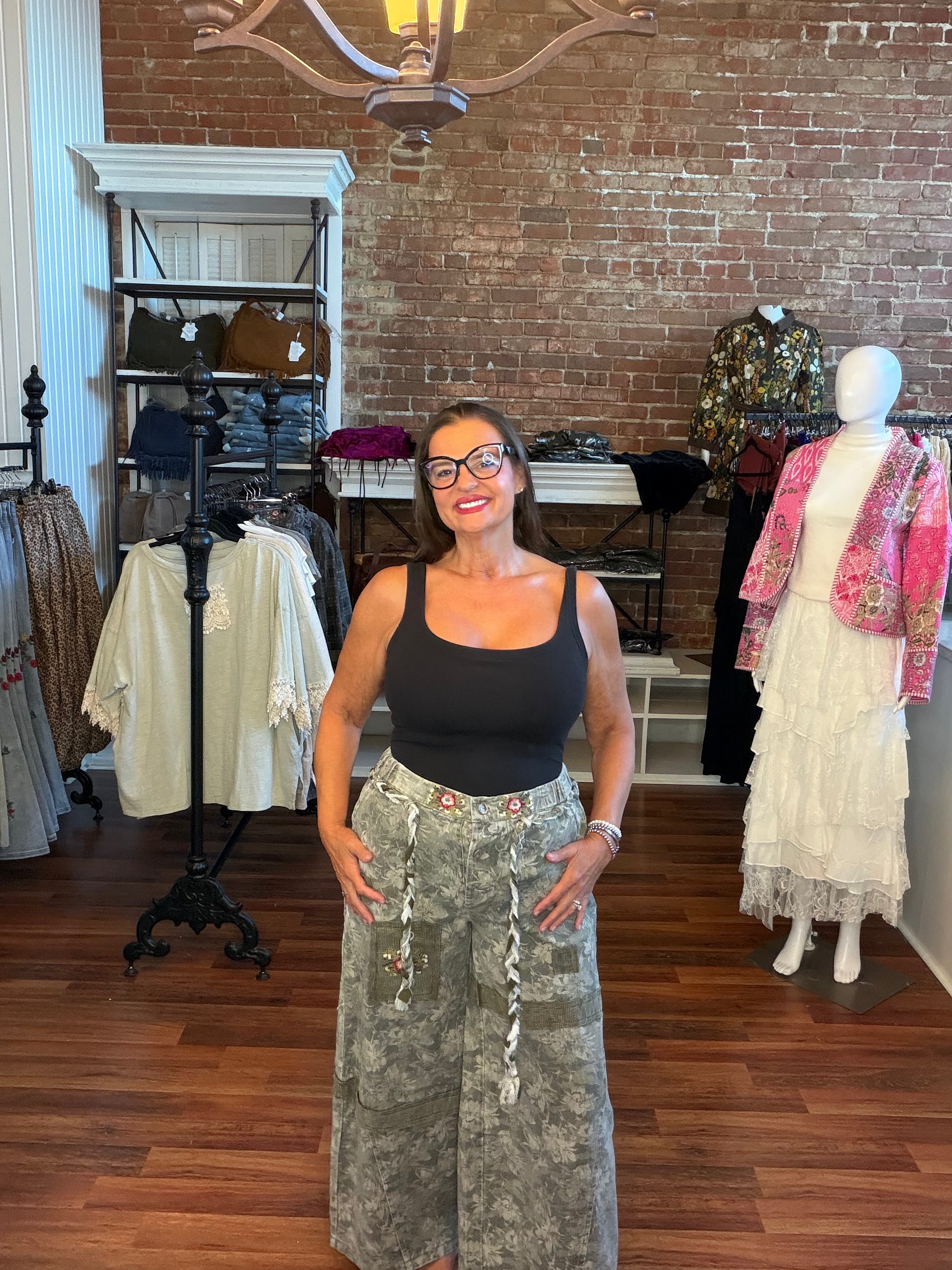 A woman wearing a black tank top and a long gray camouflage skirt stands in a boutique shop with a brick wall background, surrounded by clothing racks and mannequins.