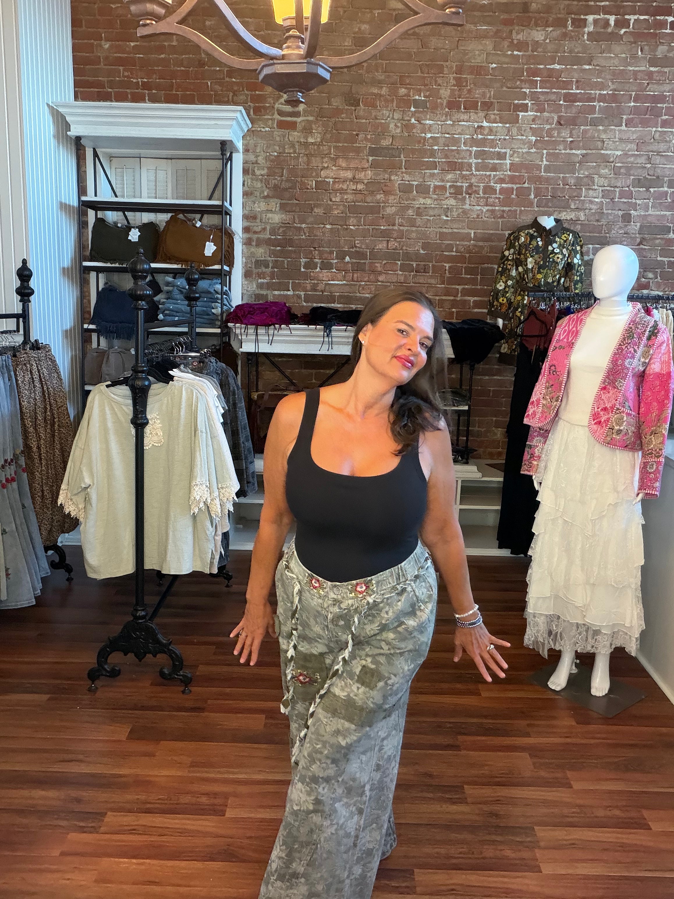 A woman posing in a boutique wearing a black tank top and a long, wide-leg skirt with a green and white camouflage pattern. The shop features a brick wall, wooden floors, and other clothing displays in the background.
