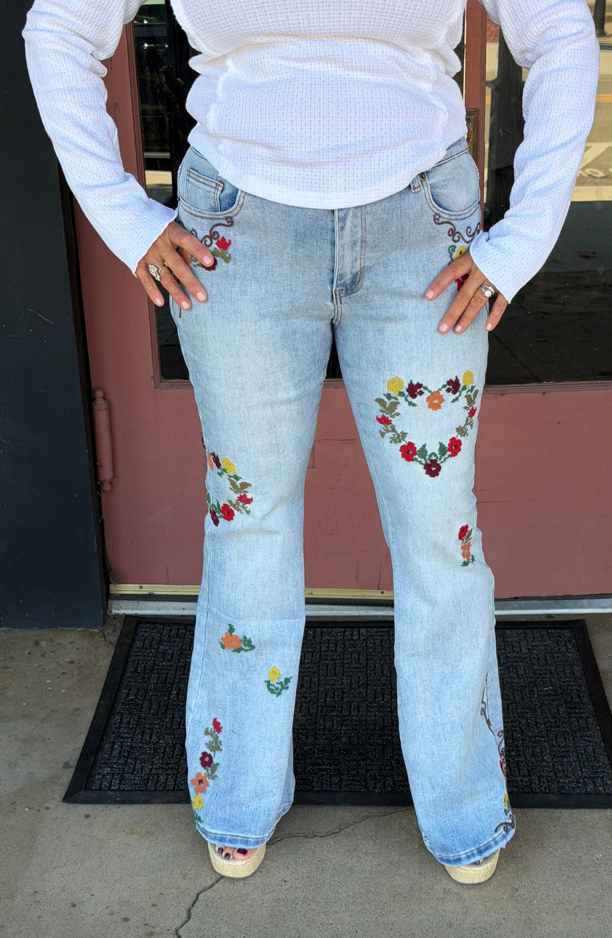 A person wearing light-wash blue flare jeans with colorful floral embroidery on the thighs and knees, paired with a white long-sleeve top, standing in front of a pink door.