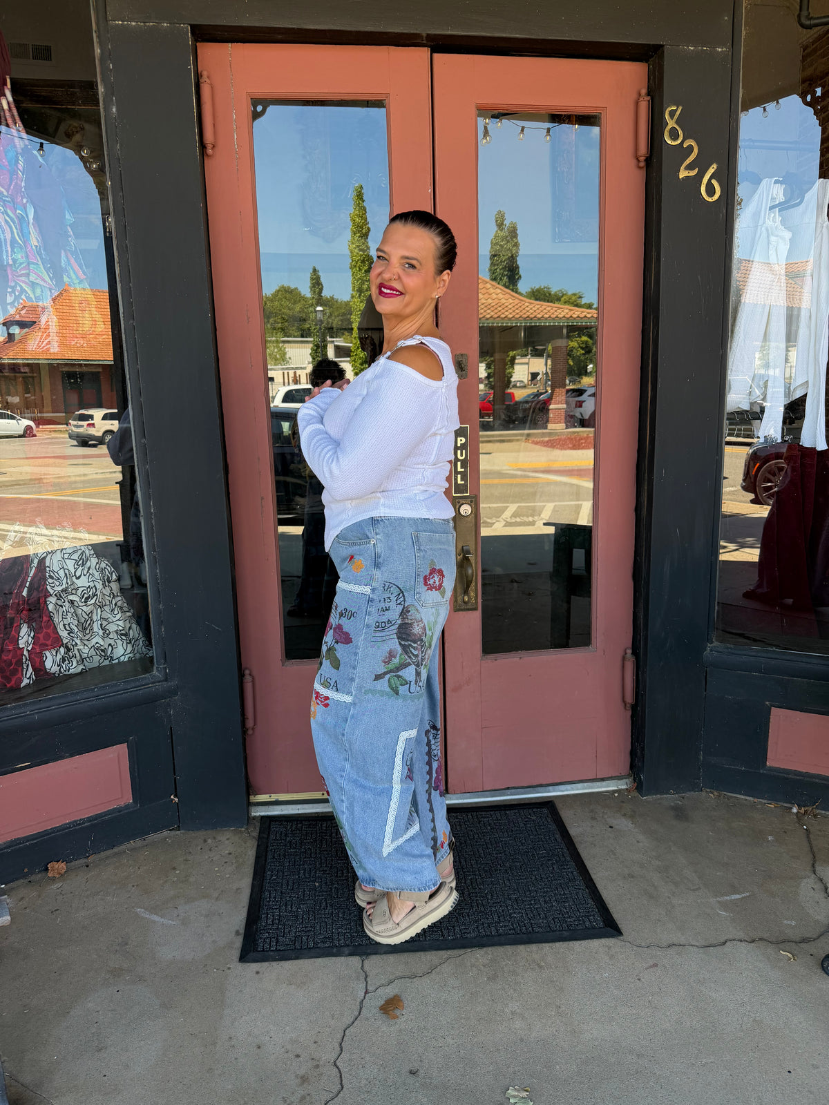 Side view Woman smiling in Alissa Patchwork Pant in light blue denim with floral and stamp patchwork, full body front view styled with rust orange boho blouse and beige heeled booties, boutique storefront