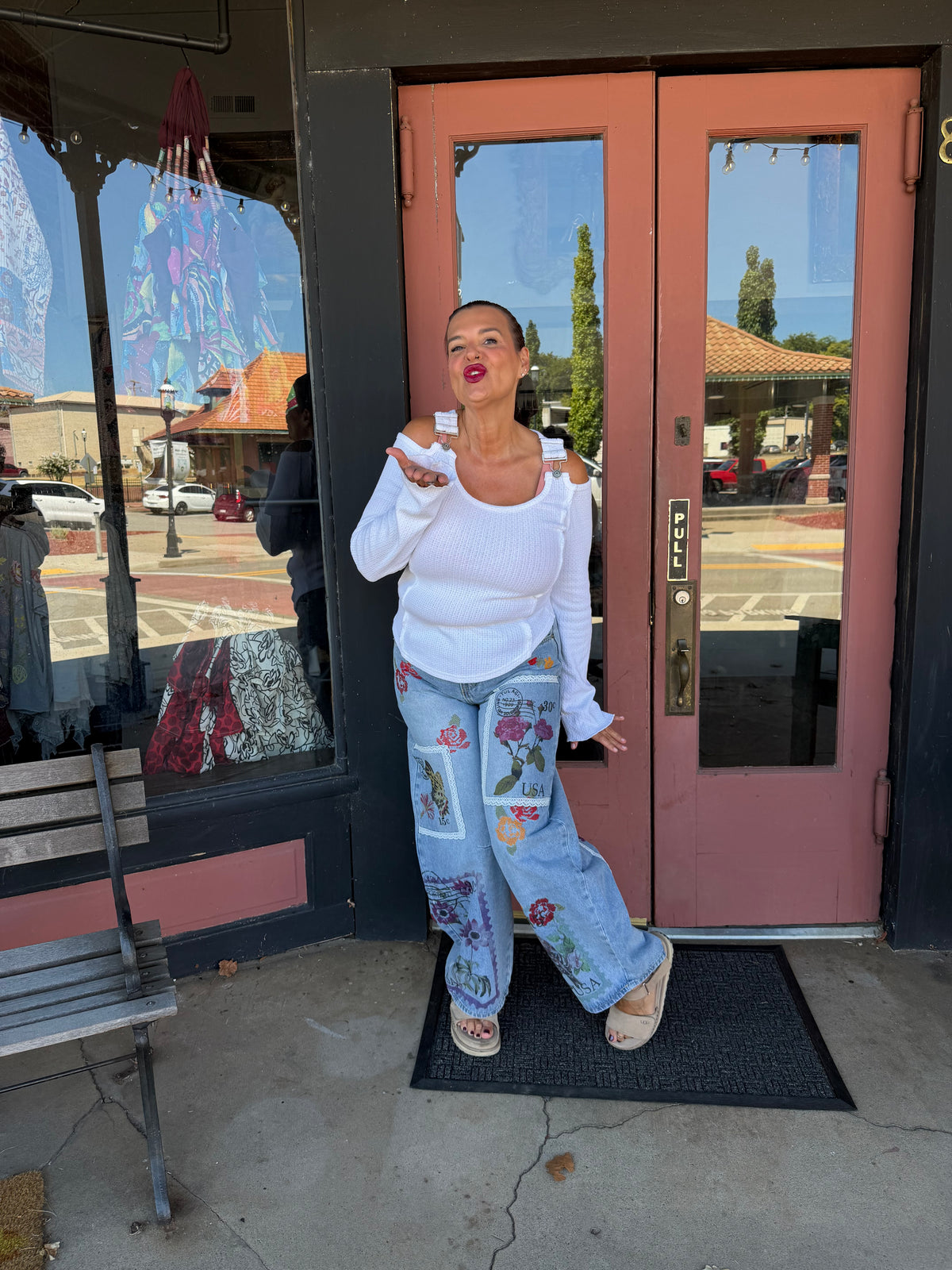A woman posing in front of a storefront with pink double doors, wearing a white long-sleeve top, light blue patchwork jeans with floral embroidery, and beige flat sandals.
