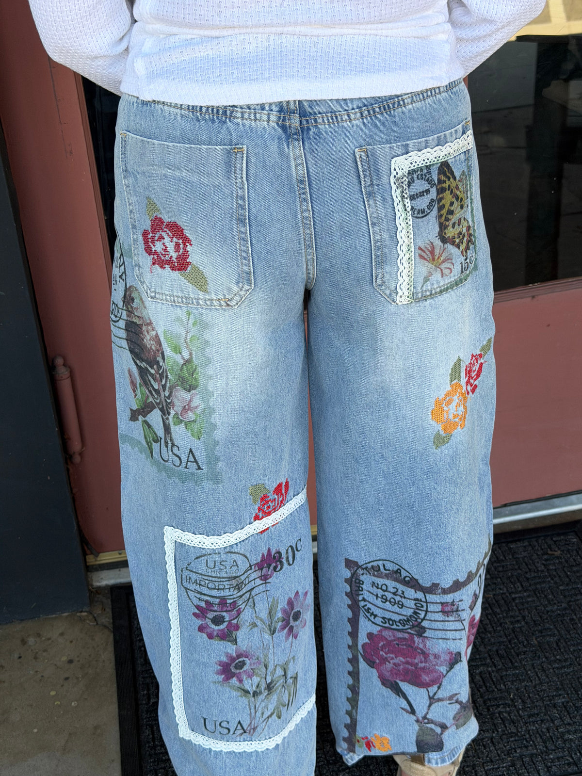 Close-up of Alissa Patchwork Pant back pockets and leg panels featuring colorful butterfly, rose, and vintage postage stamp embroidery on light blue denim Back view