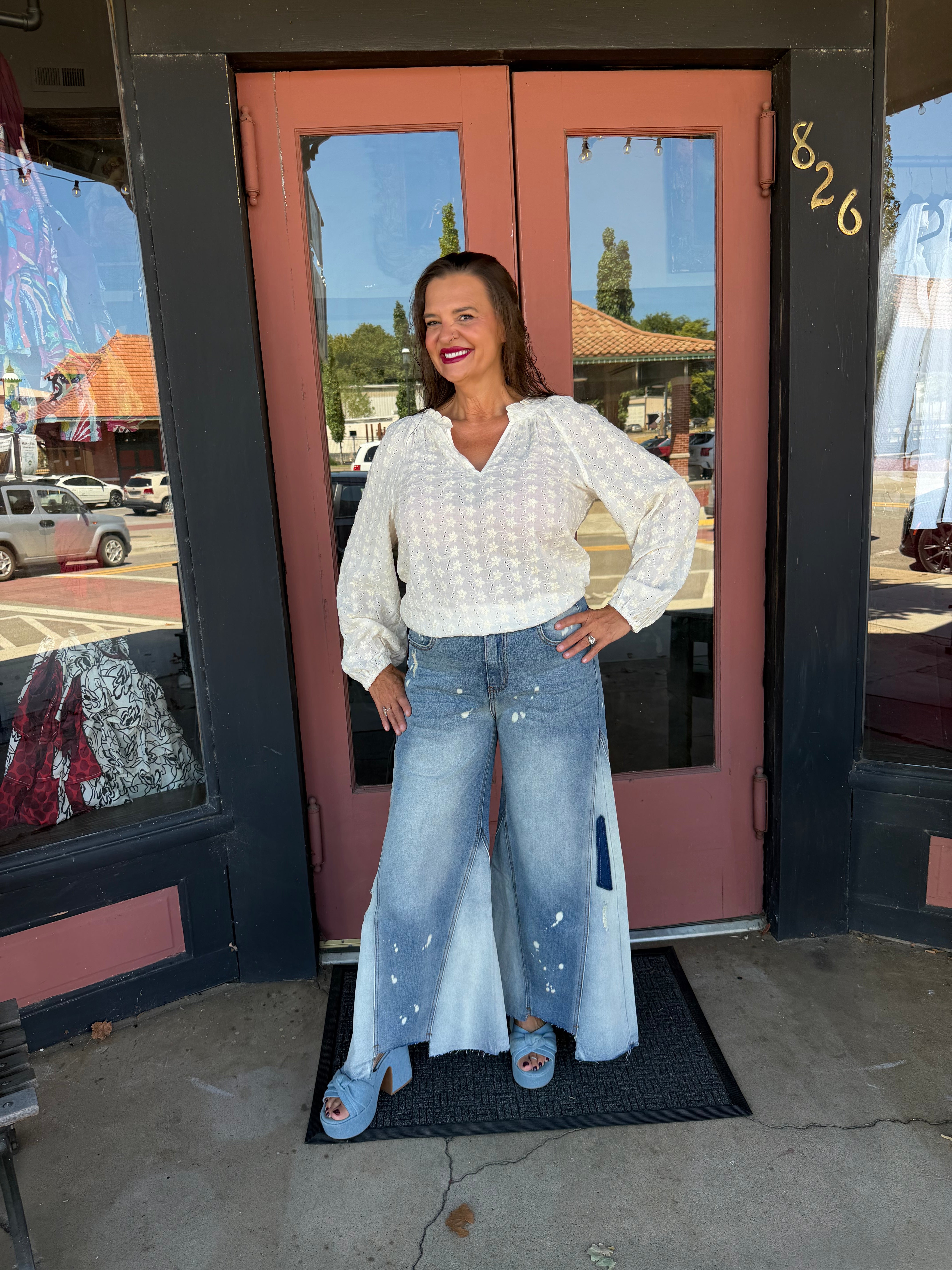 Woman smiling in a white floral eyelet long sleeve blouse with ruffle V-neckline, styled with wide-leg paint-splatter jeans and blue platform heels, full body front view outdoors