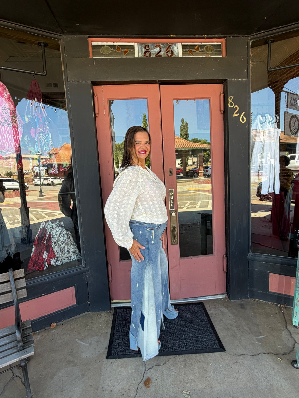 Woman smiling in Jolene Wide Leg Flare Jeans with paint splatter distressing, contrast patchwork godet panels, and raw frayed hem, full body side view styled with white eyelet blouse, boutique exterior