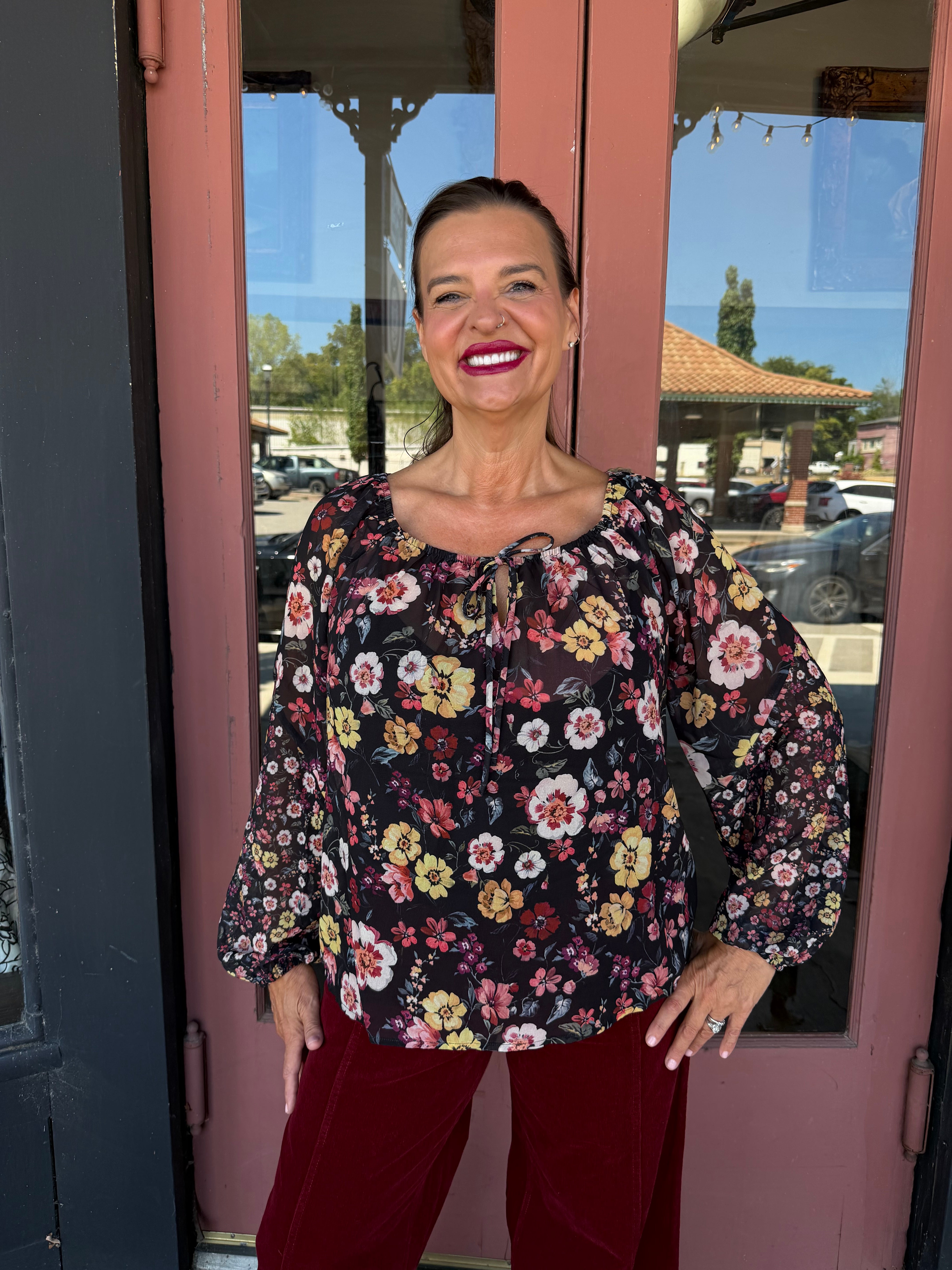 A woman stands in front of a pink storefront door wearing a black long-sleeve blouse with a colorful floral print and burgundy pants.