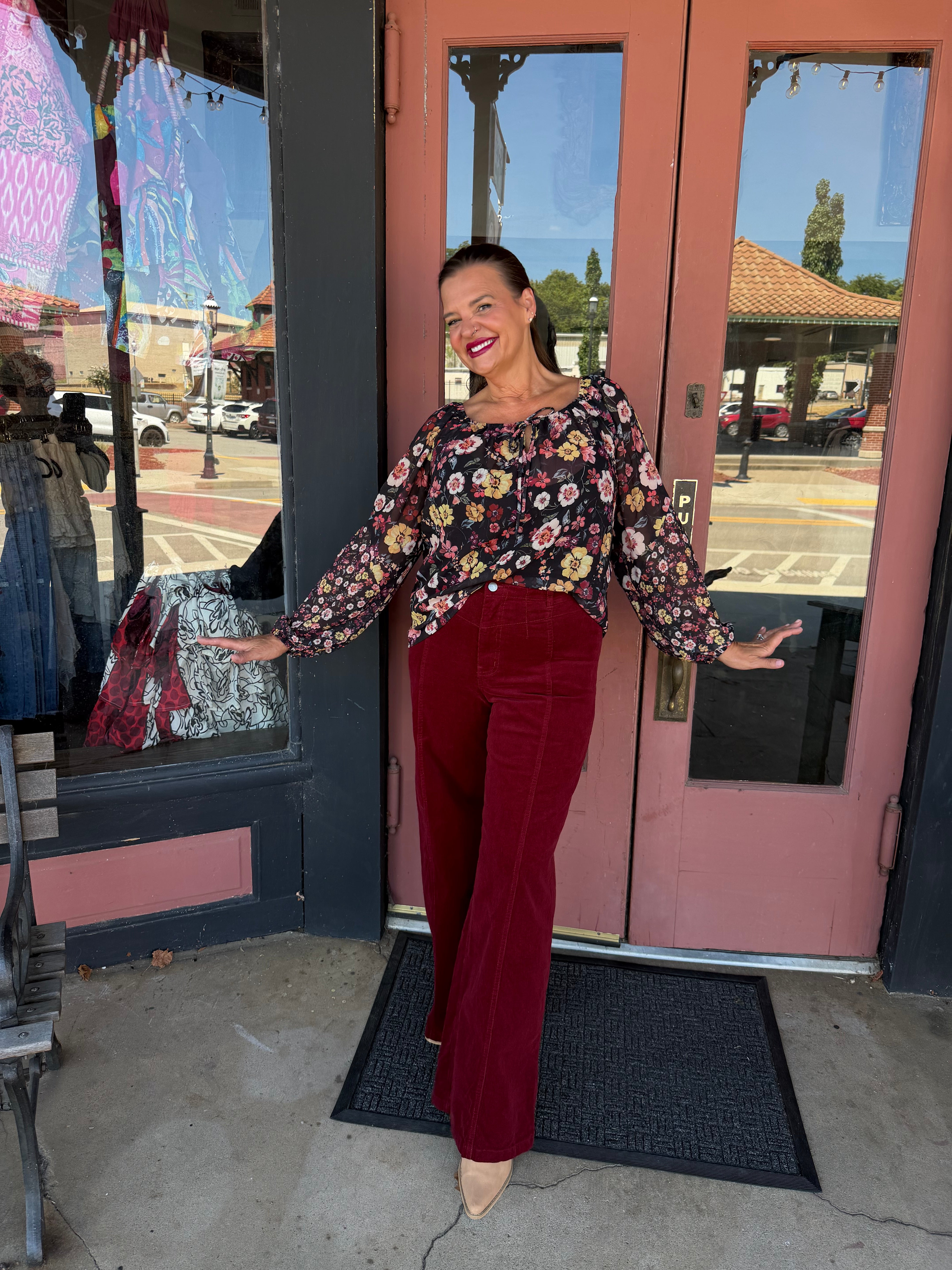 A woman posing in front of a storefront with pink doors, wearing a long-sleeve floral print top and burgundy velvet flare pants.