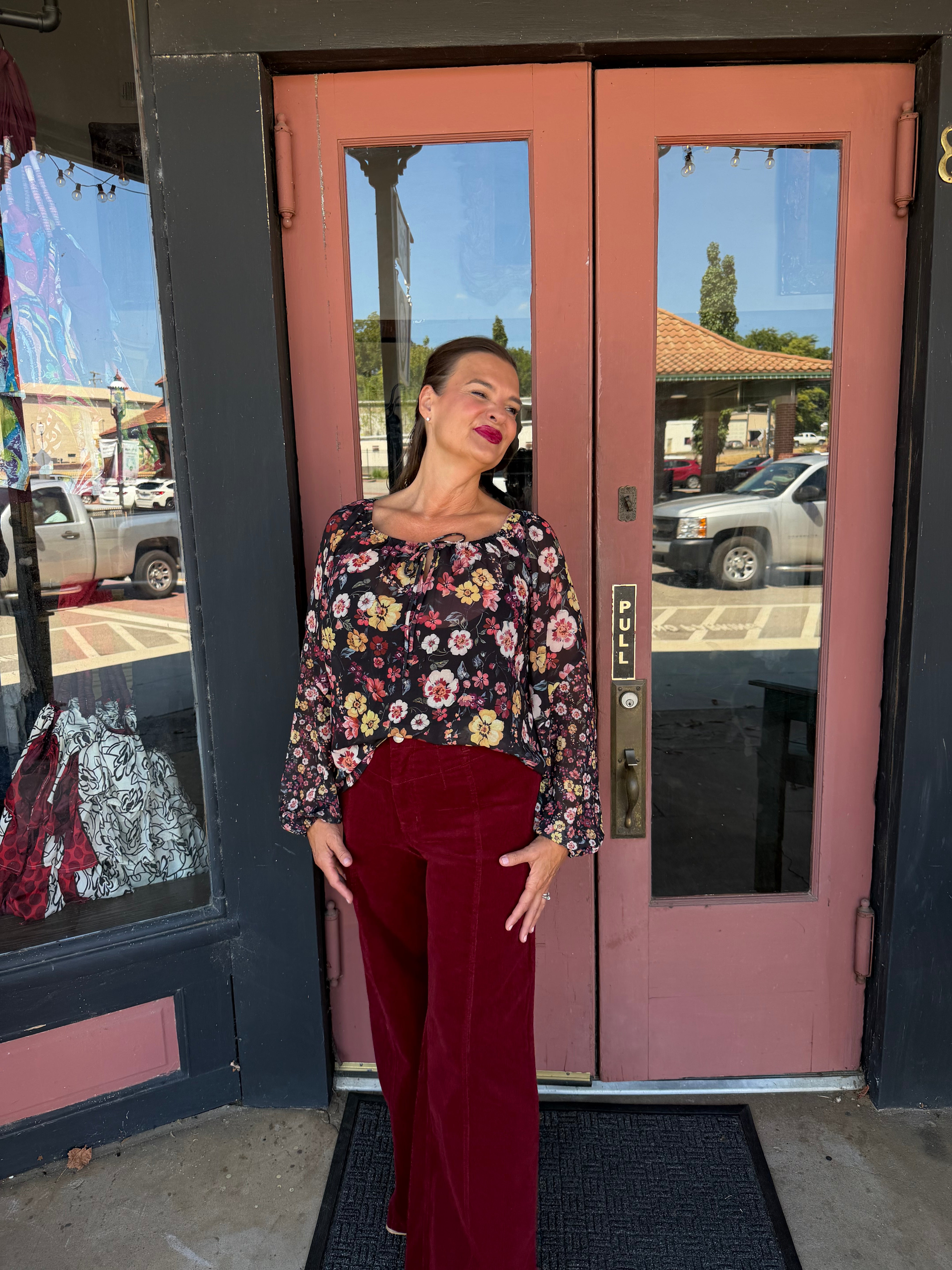 A woman stands in front of a pink storefront door wearing a floral long-sleeve top and burgundy wide-leg pants.