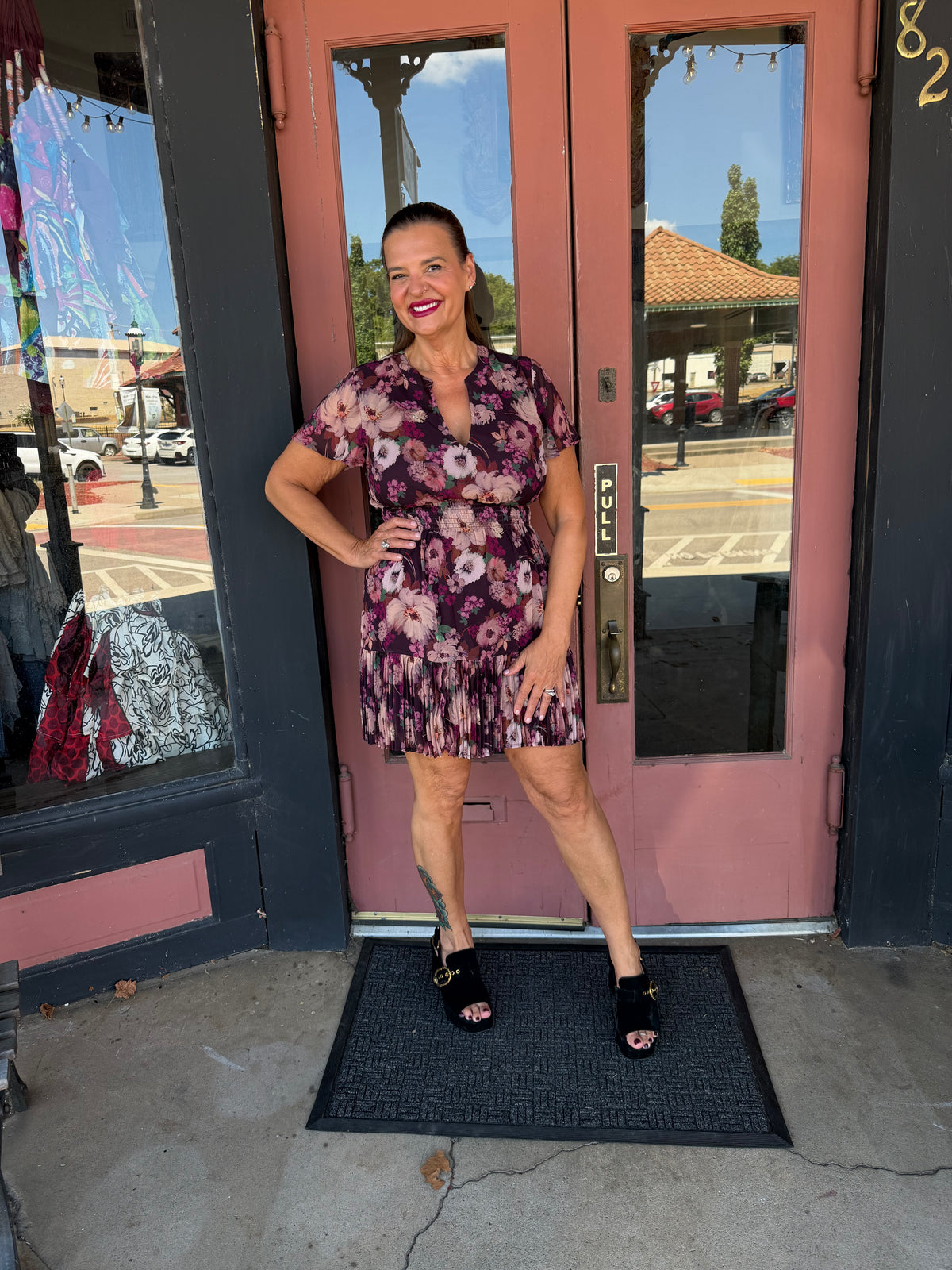 A woman stands wearing a short-sleeve floral mini dress with a pink, purple, and white pattern, paired with black slide sandals.