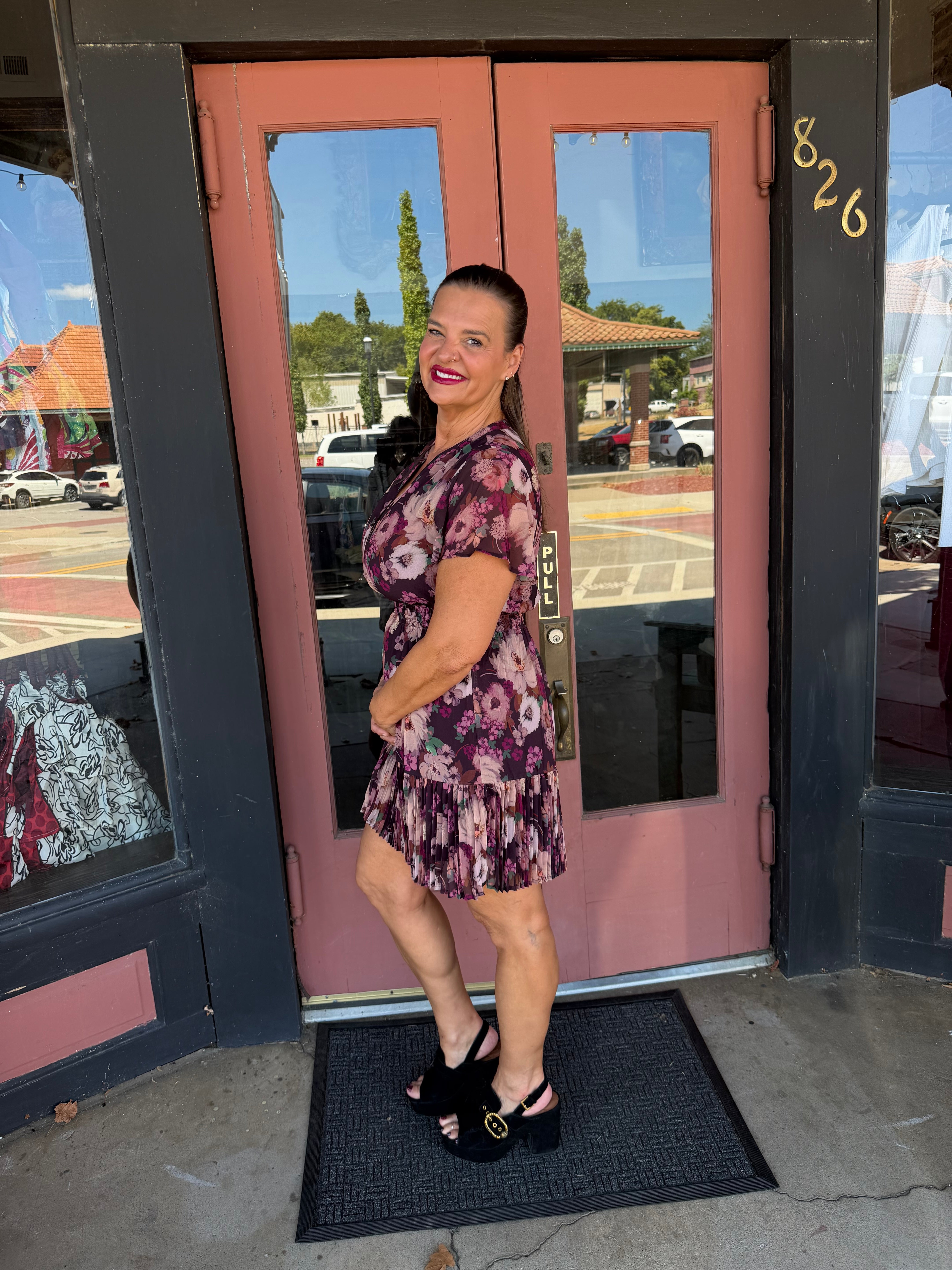 A woman wearing a short-sleeve floral mini dress with a purple, pink, and black pattern, paired with black platform heels.