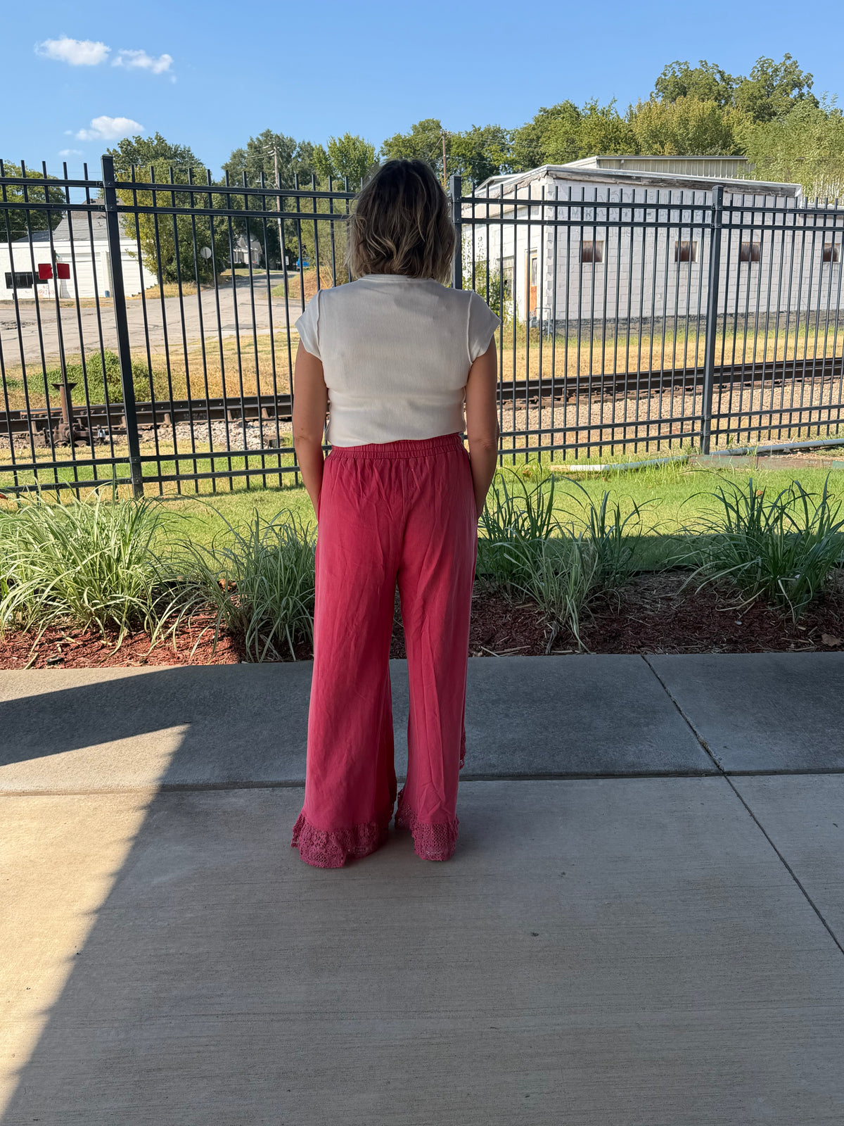 A woman stands with her back to the camera on a concrete patio, wearing a cream short-sleeve top and bright pink wide-leg pants with ruffle trim at the hem. In the background is a black metal fence, green plants, and a white building under a blue sky.