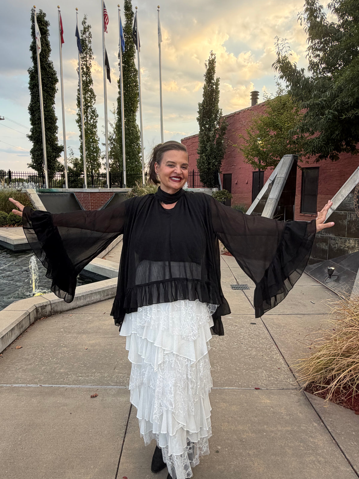 A woman stands outdoors with her arms outstretched, wearing a sheer black long-sleeve top layered over a white ruffled tiered skirt. The background shows a brick building, trees, flagpoles, and a cloudy sky.