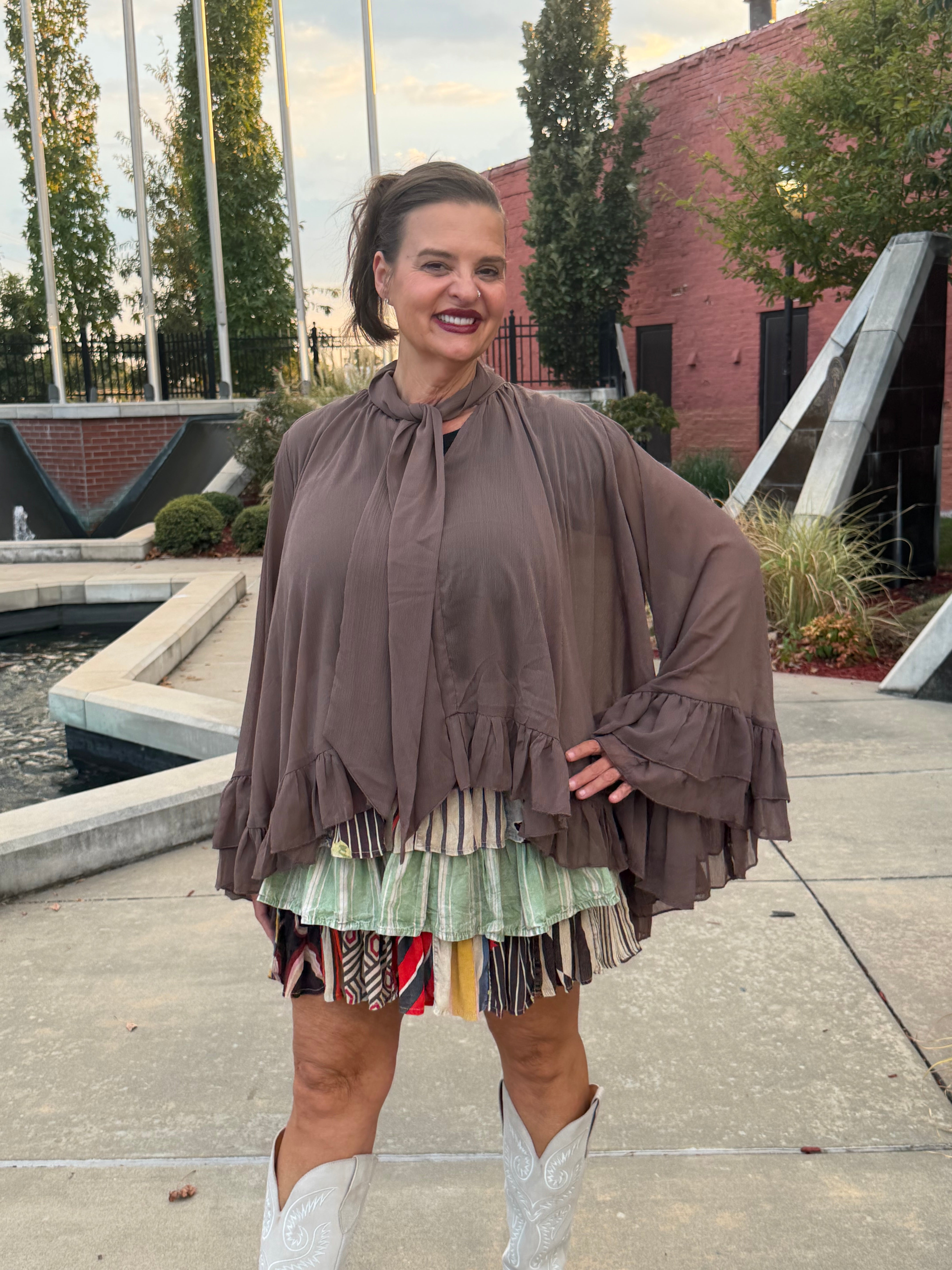 A woman wearing a brown tiered long-sleeve blouse and a multicolor striped fringe skirt, standing outdoors on a concrete plaza with a brick building and trees in the background.