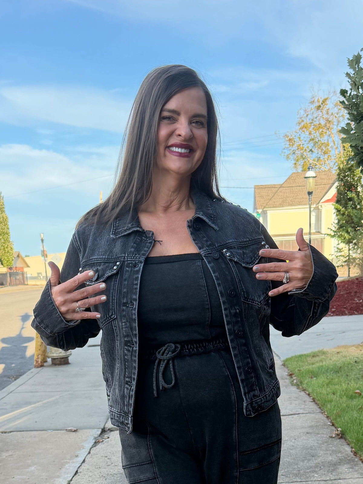 A woman wearing a black denim jacket, black top, and black wide-leg pants walks on a sidewalk outdoors, gesturing with her hands. The background shows a street, grass, trees, and a blue sky.