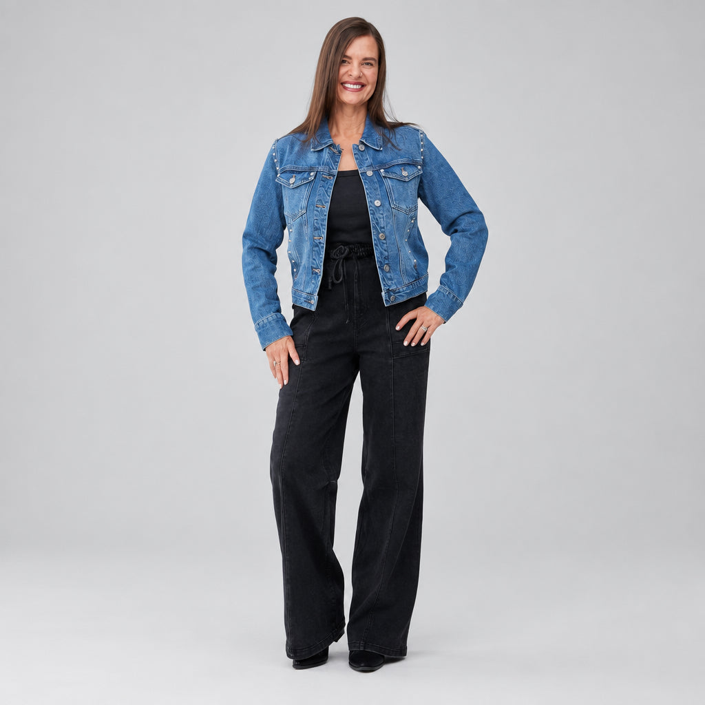 A woman stands wearing a blue denim jacket over a black top and black wide-leg pants.
