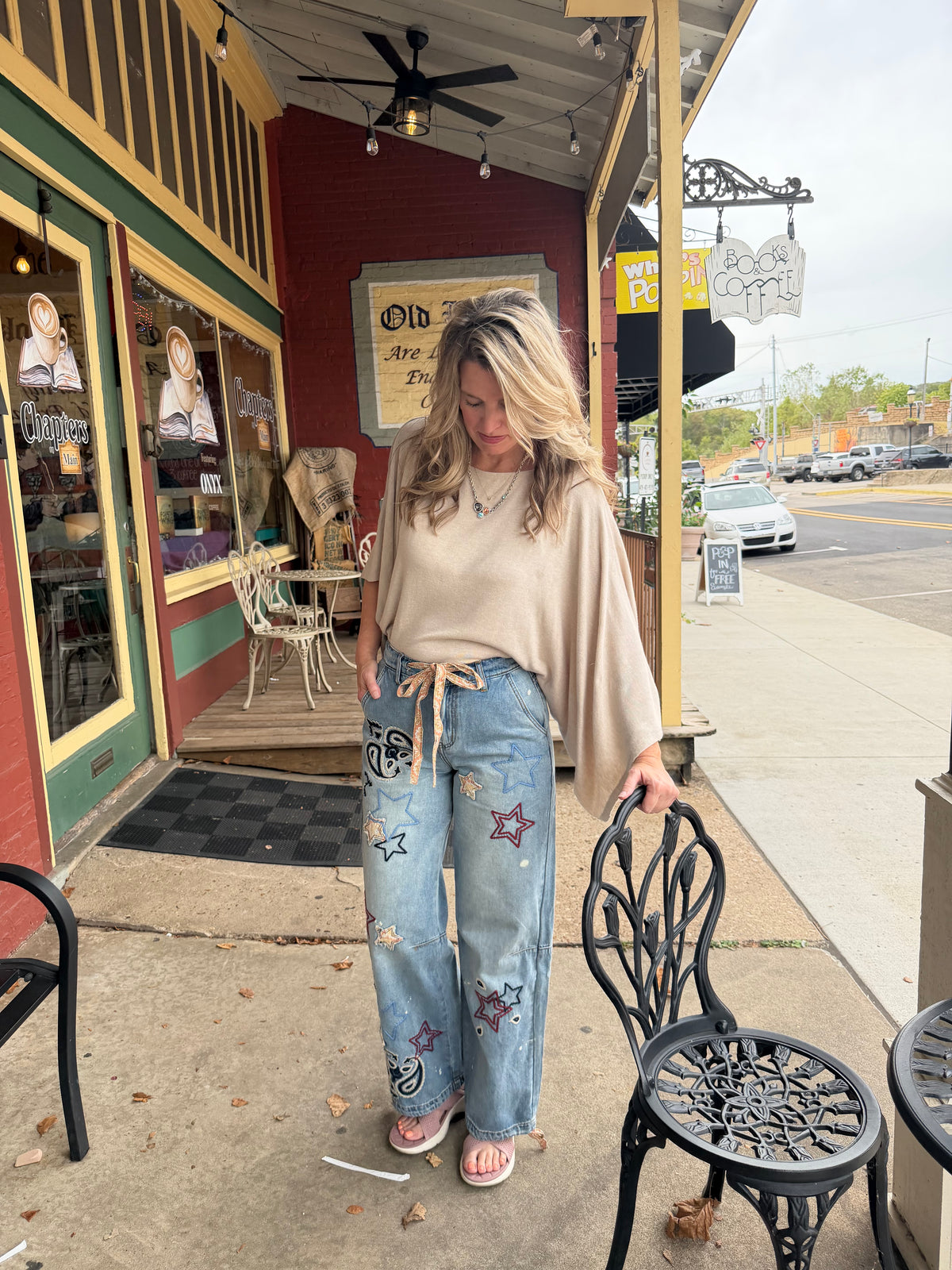 A woman stands on a sidewalk in front of a storefront wearing a beige long-sleeve tunic top and light-wash blue jeans with star and floral patches. She is holding onto a black metal chair beside the store entrance.