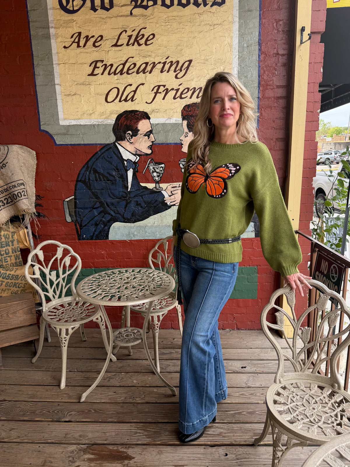 A woman stands on a wooden porch wearing a green knit sweater with a large orange butterfly graphic on the front, paired with blue jeans. The background features a rustic brick wall with a painted mural and white metal patio furniture.