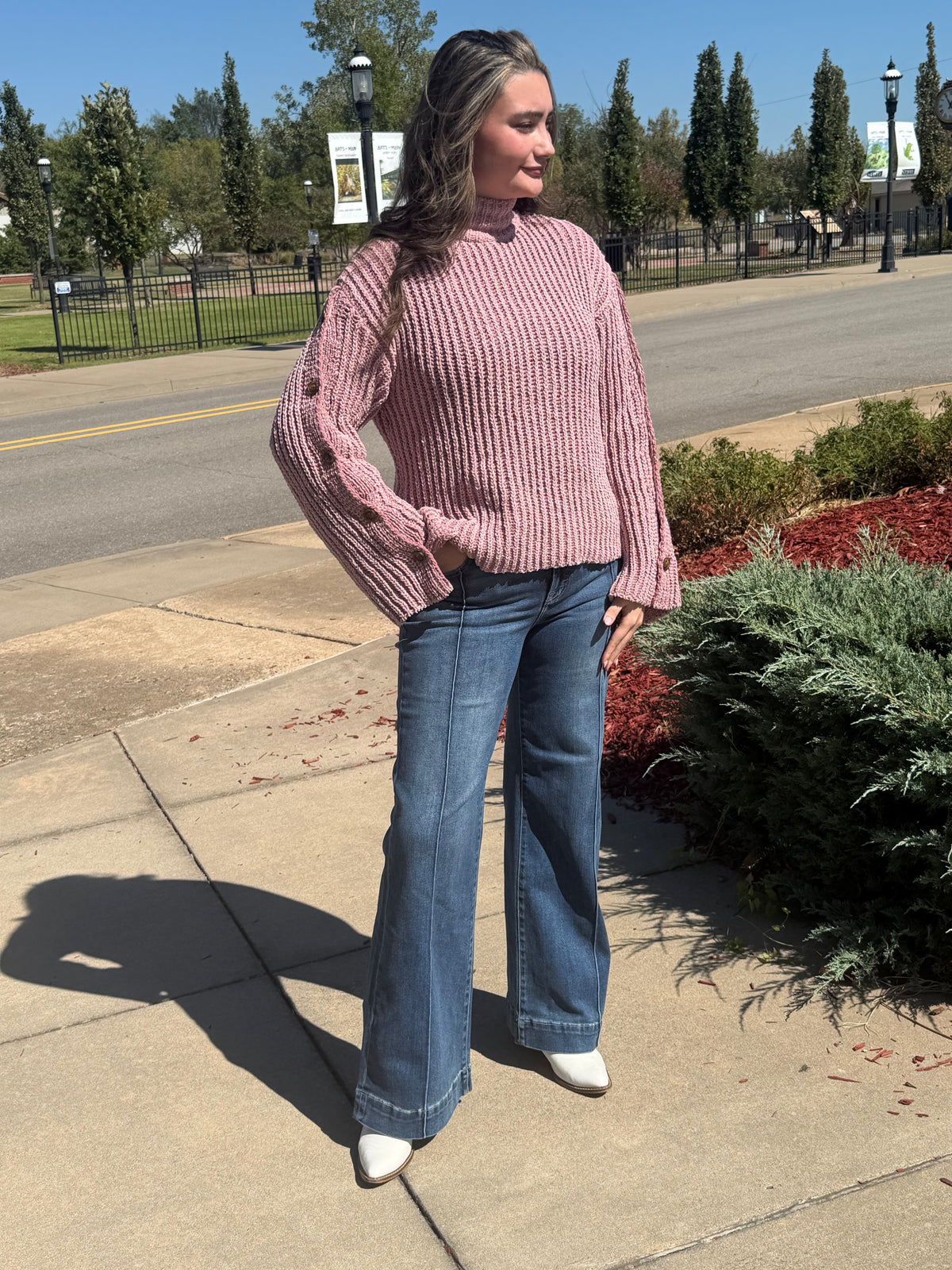 Angled view of a model wearing a dusty mauve mock-neck sweater, showing sleeve buttons and flared cuffs, paired with jeans