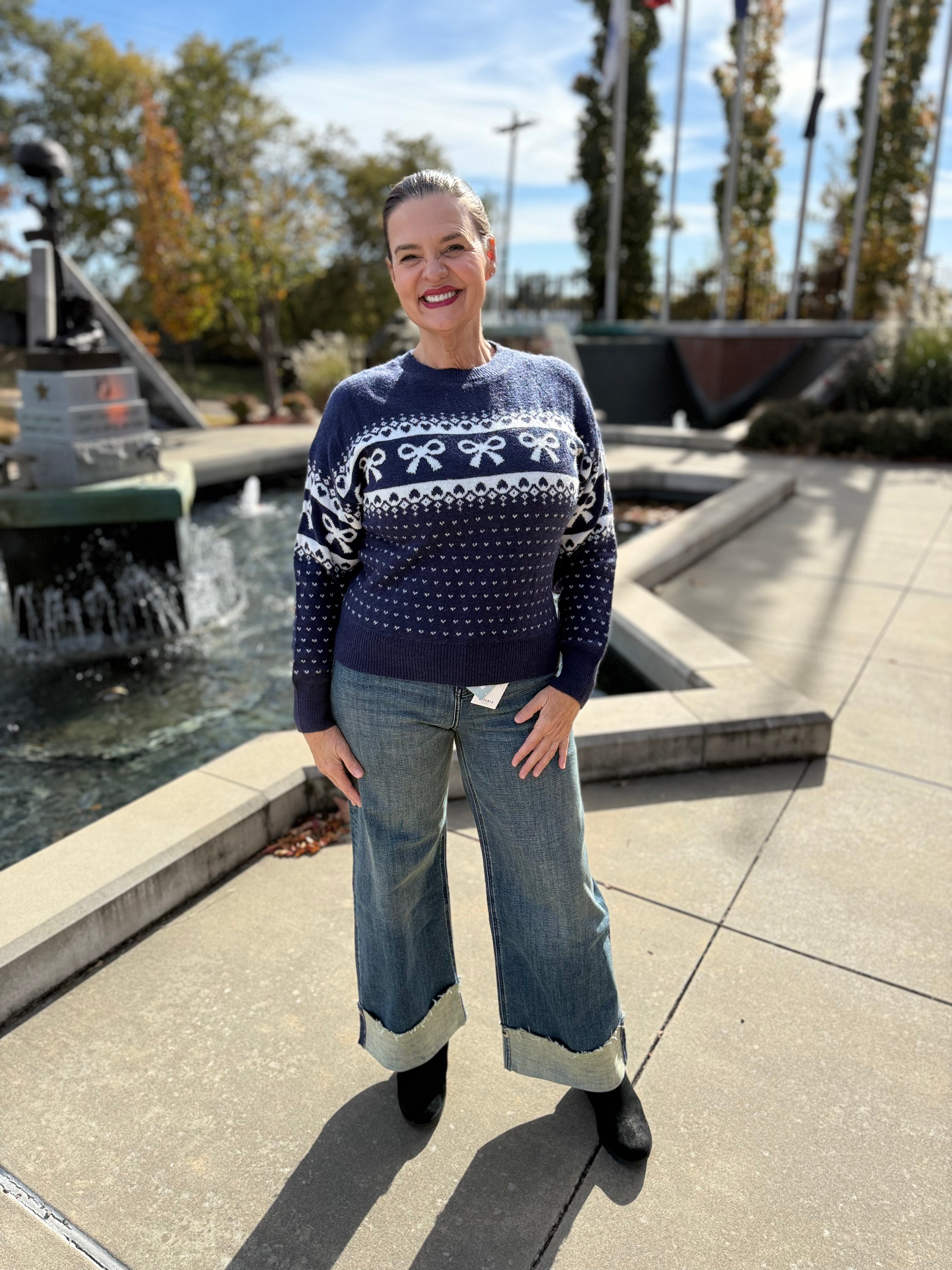Woman wearing a blue sweater with white patterns and jeans standing outdoors near a water feature.