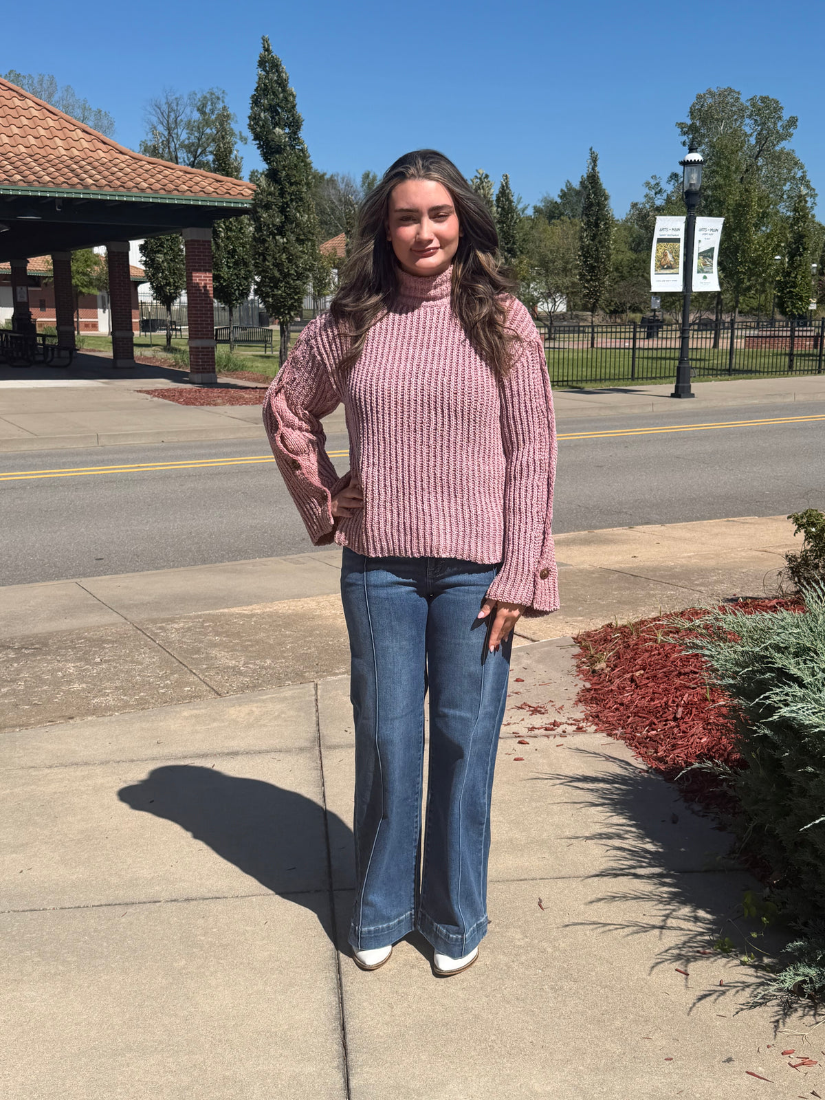 Front view of a dusty mauve ribbed mock-neck sweater with wide button-detail sleeves, modeled with blue jeans and white boots.