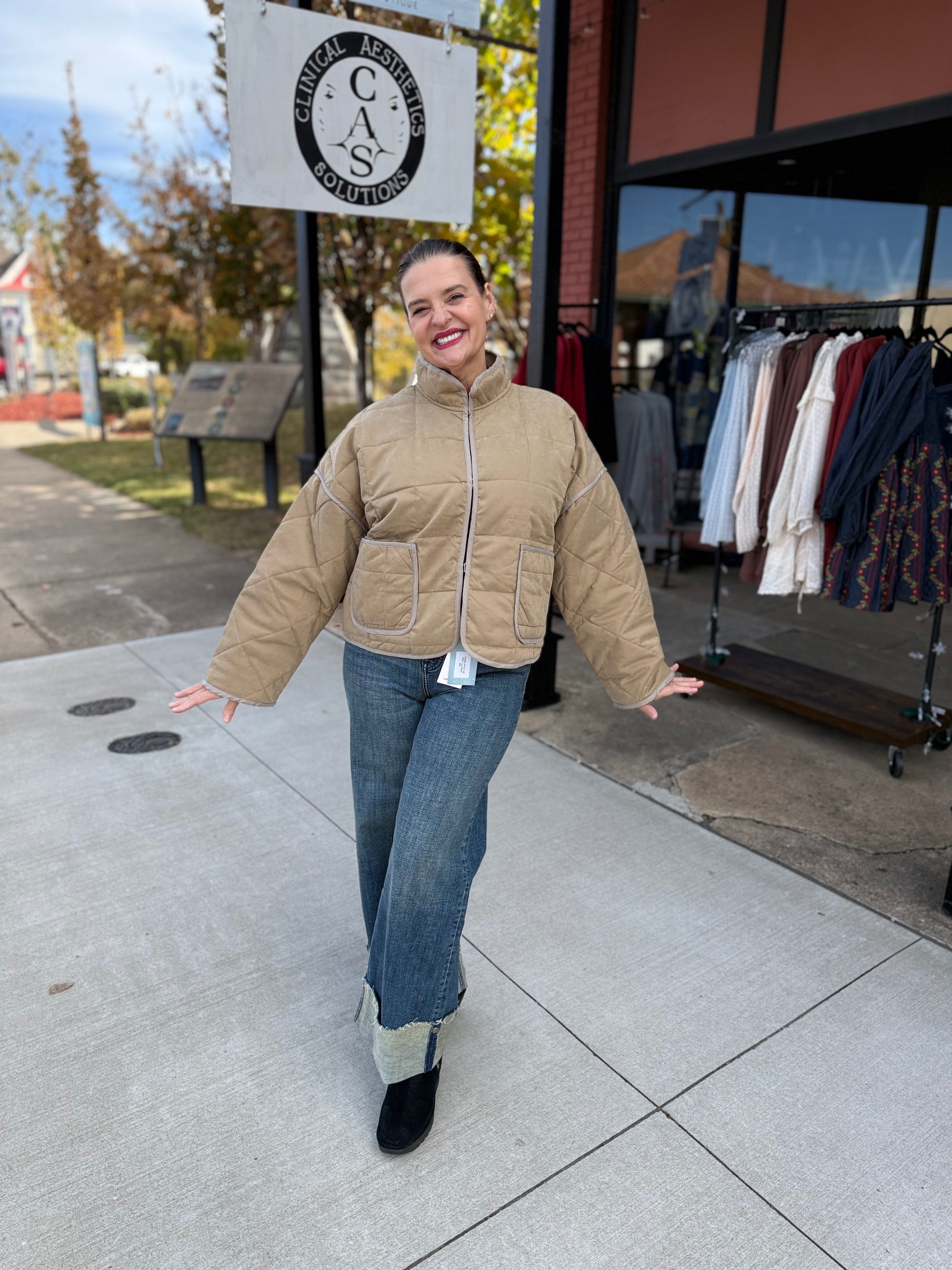 Woman in a beige puffer jacket and blue jeans standing on a sidewalk with a store in the background.