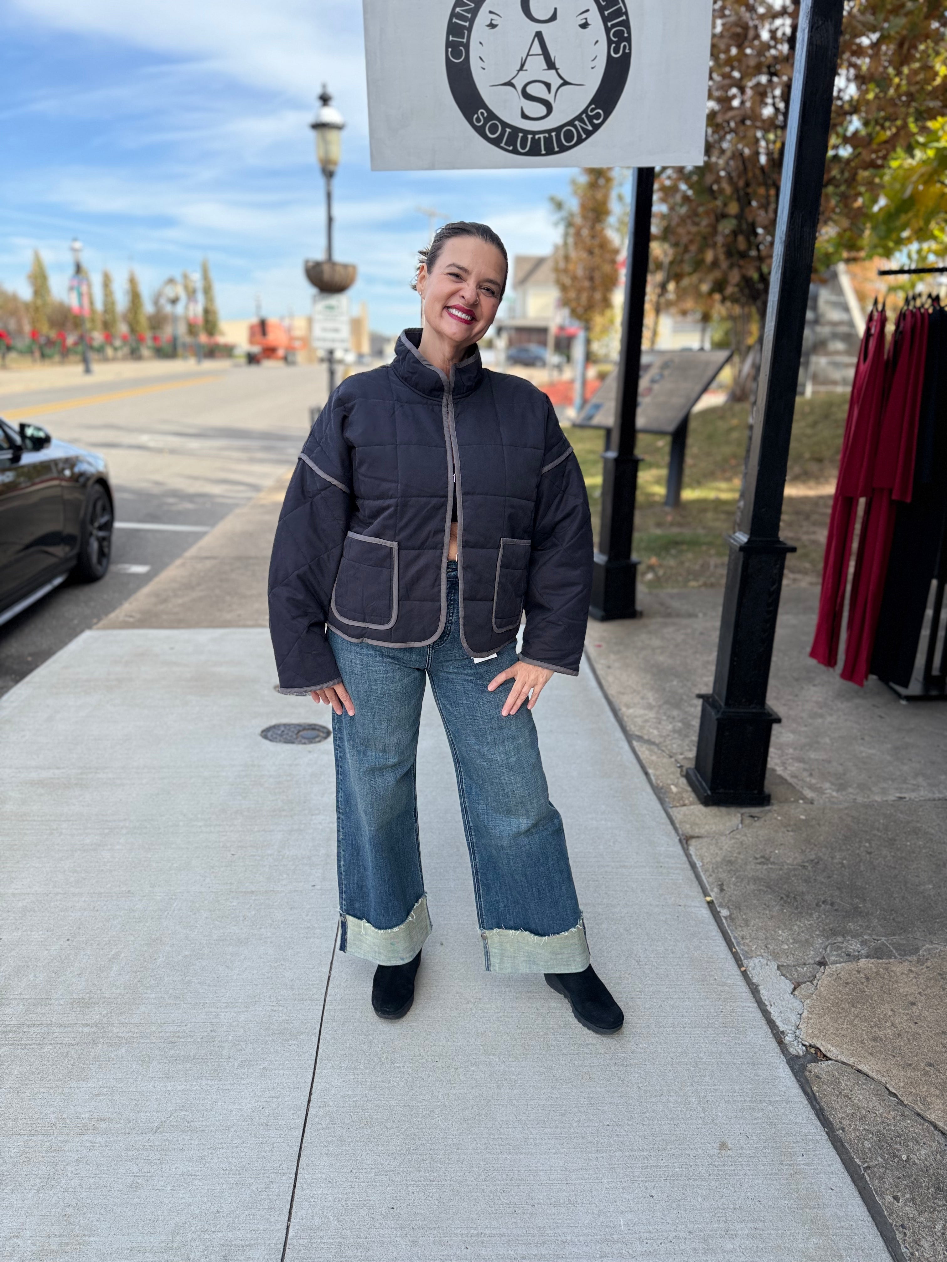Woman in a dark jacket and jeans standing on a sidewalk with a sign in the background.