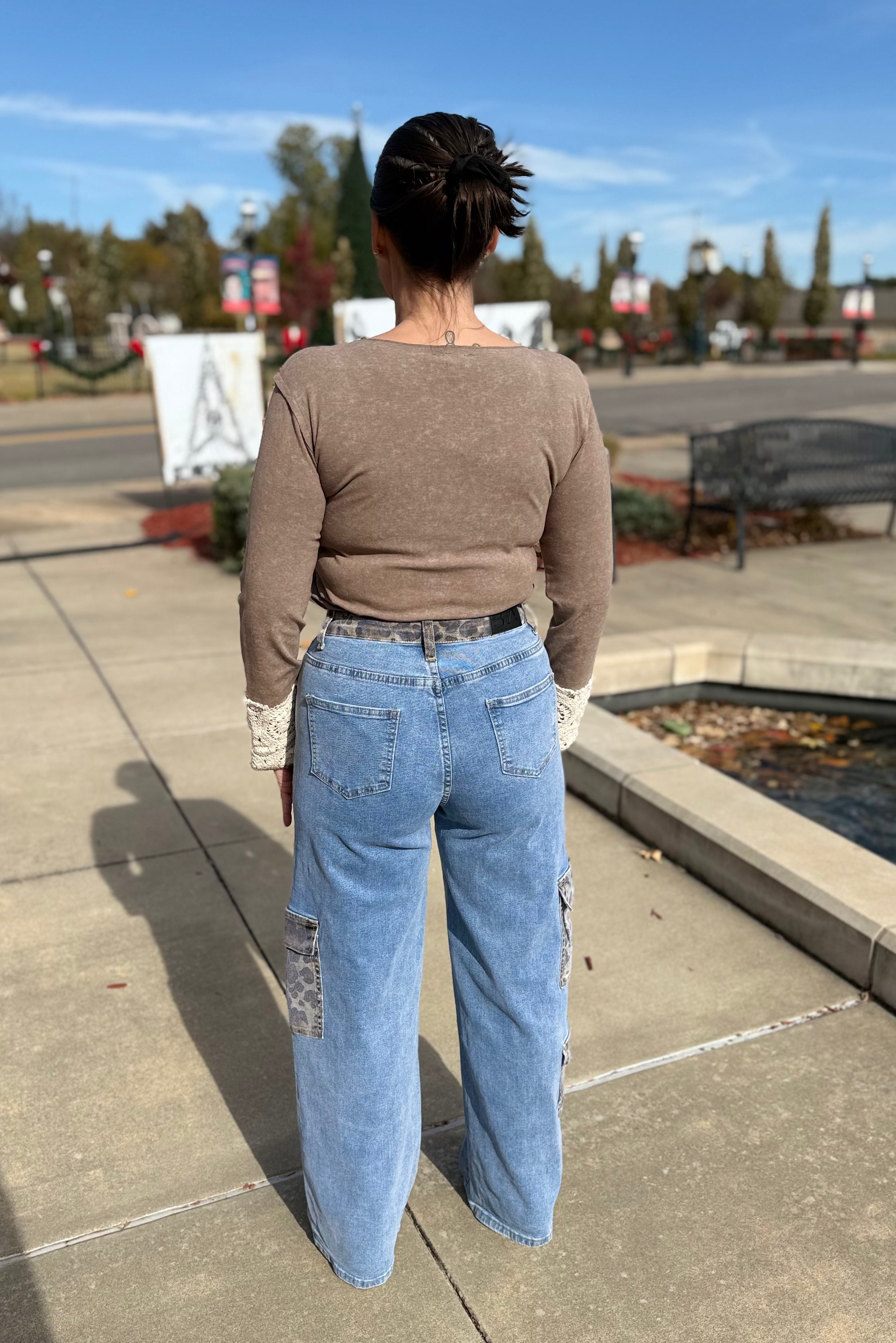 Person wearing a brown long-sleeve top and blue jeans standing on a sidewalk with trees and a bench in the background.