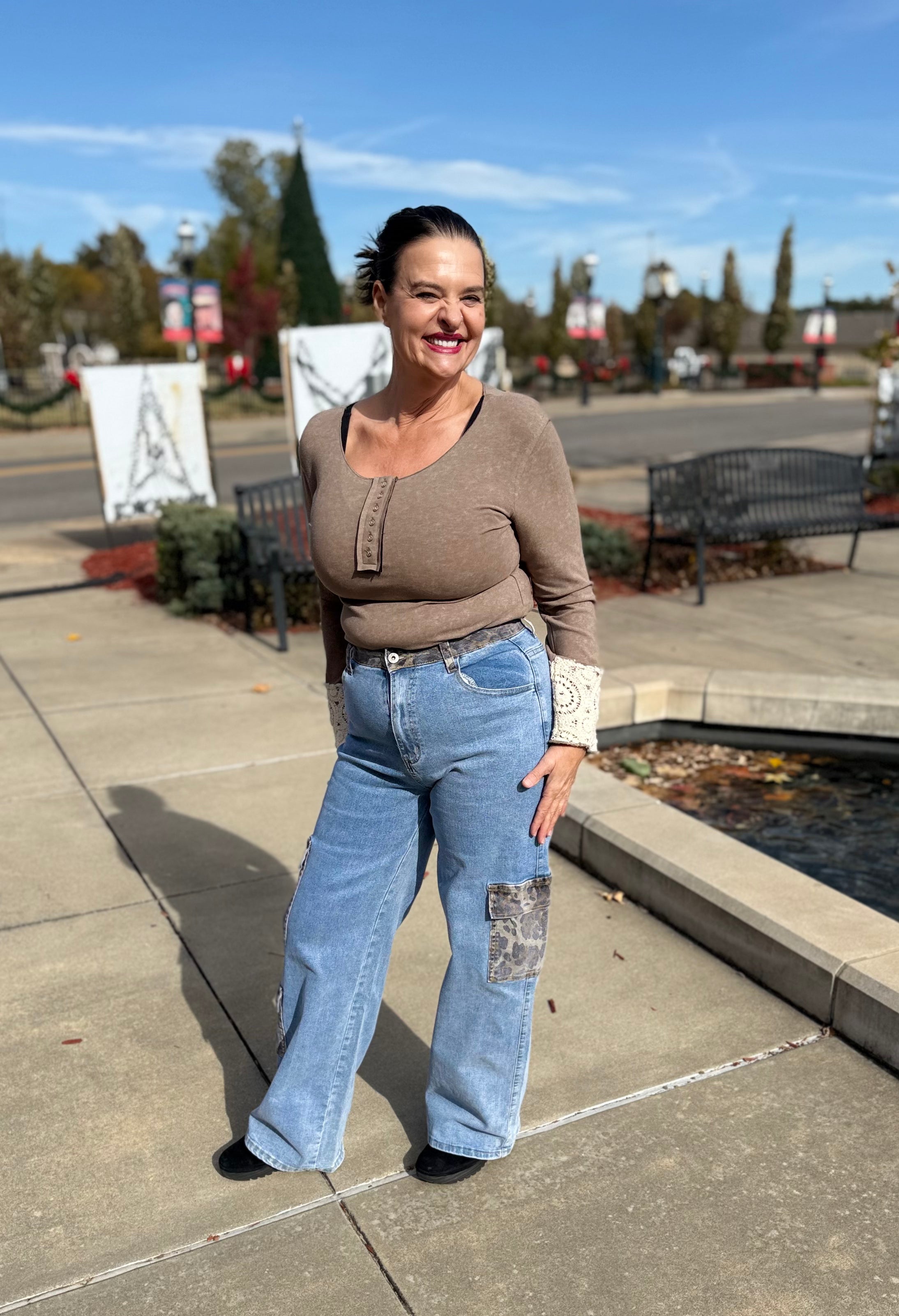 Woman in brown sweater and blue jeans standing on a sidewalk with trees and benches in the background.
