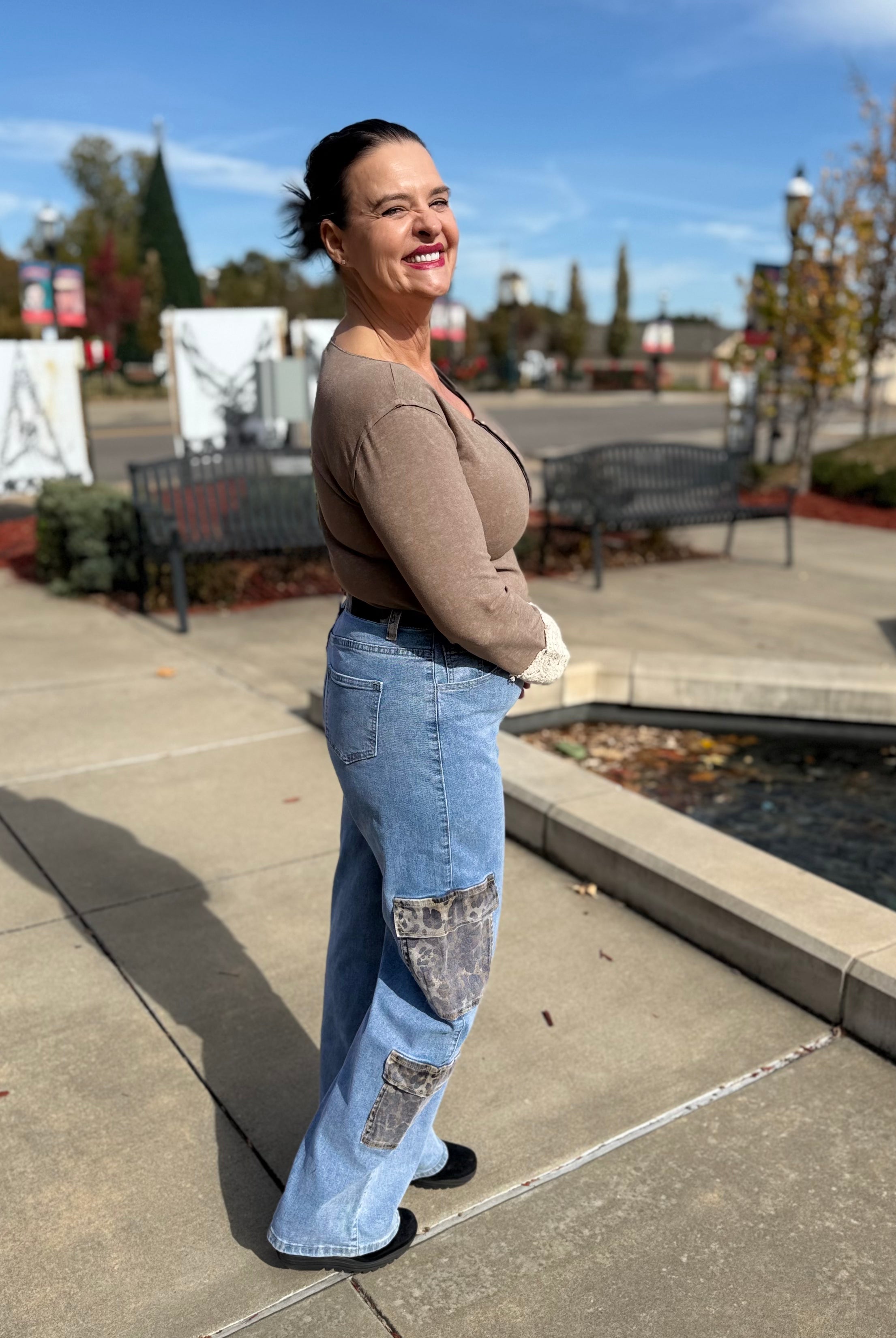 Woman standing on a sidewalk wearing a brown sweater and blue jeans with patches.