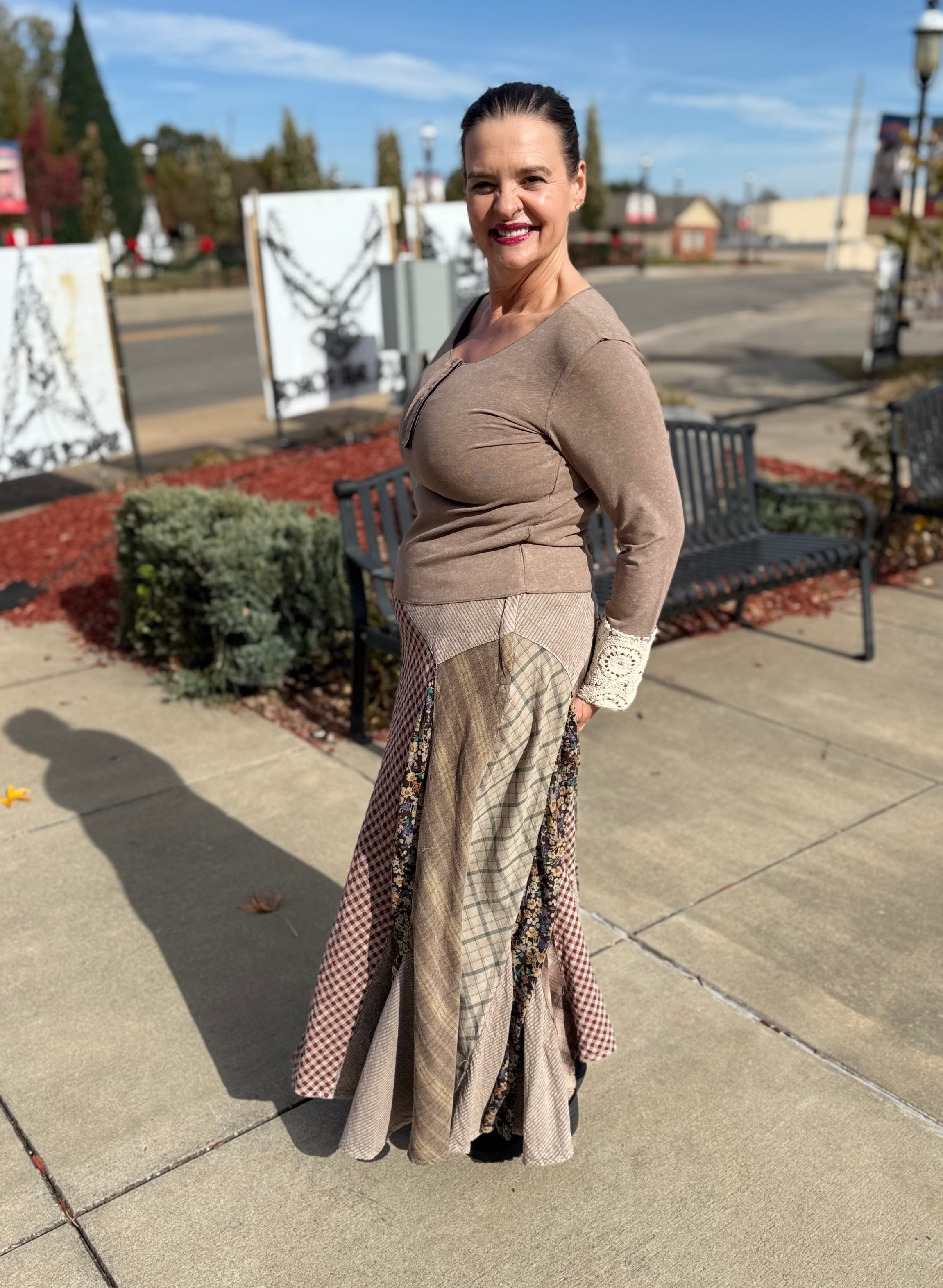 Woman in a brown top and patterned skirt standing on a sidewalk with a bench and trees in the background.