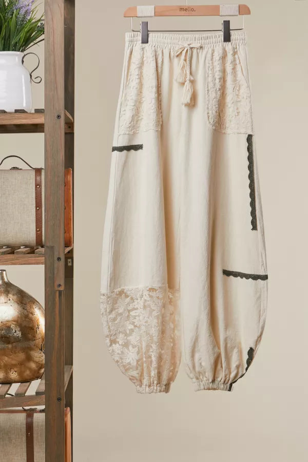 Beige pants with lace details hanging on a wooden hanger against a beige wall.