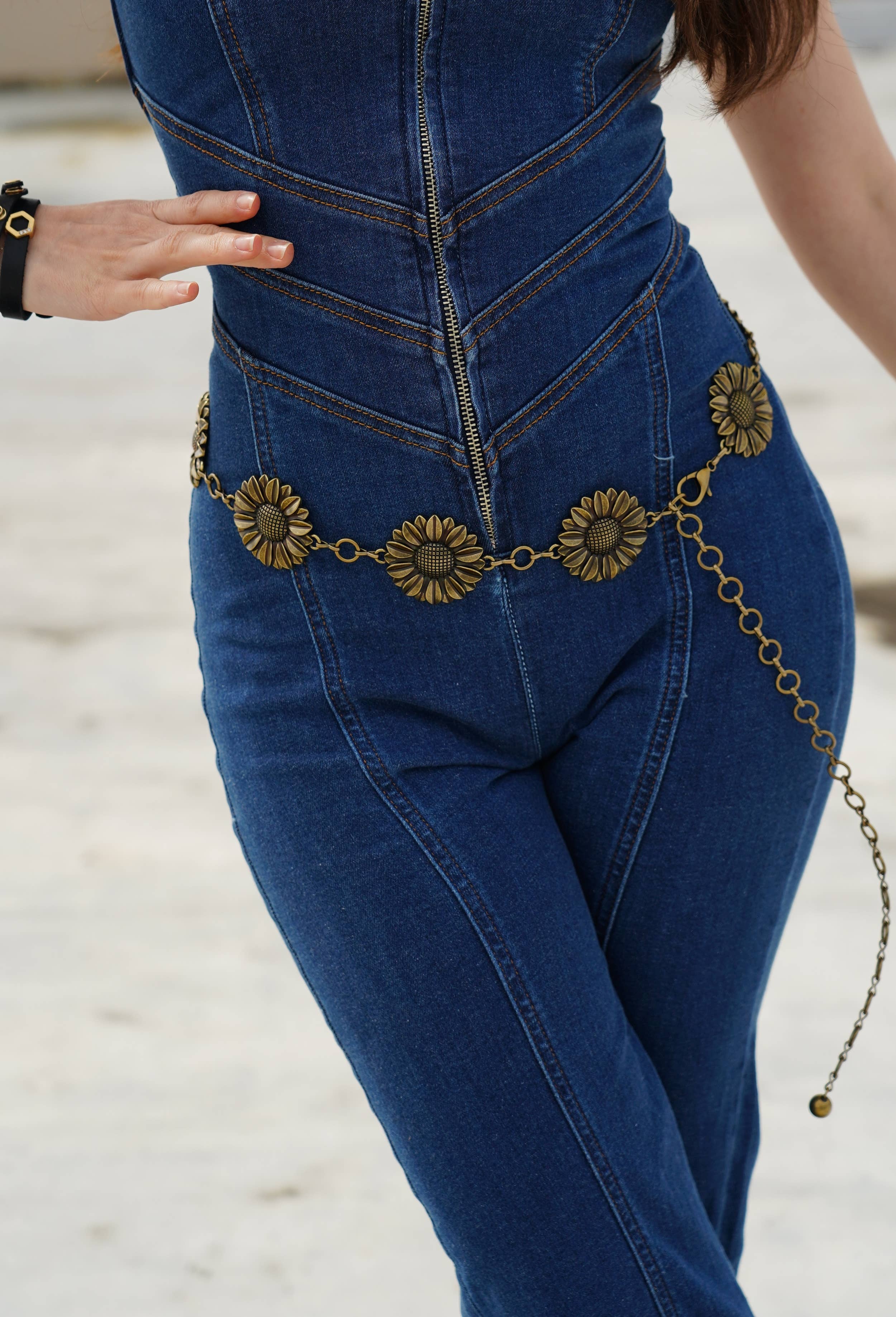 A close-up of a person wearing a blue denim jumpsuit with a decorative bronze sunflower chain belt at the waist, standing outdoors on a light sandy surface.