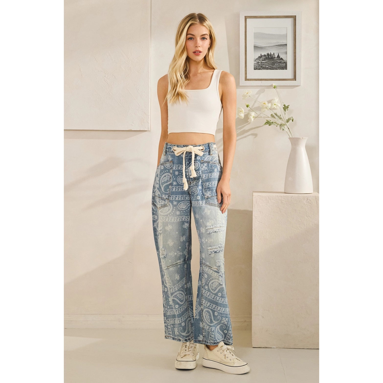 Woman wearing a white tank top and patterned blue jeans in a room with a vase and framed picture.