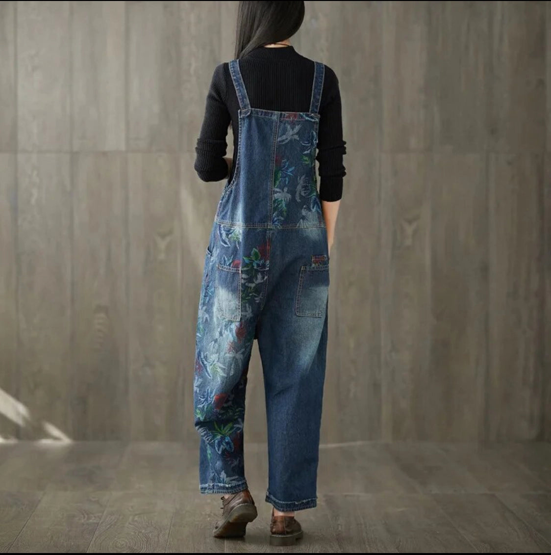 Woman wearing a pair of blue denim painted overalls rear view against a wooden wall.