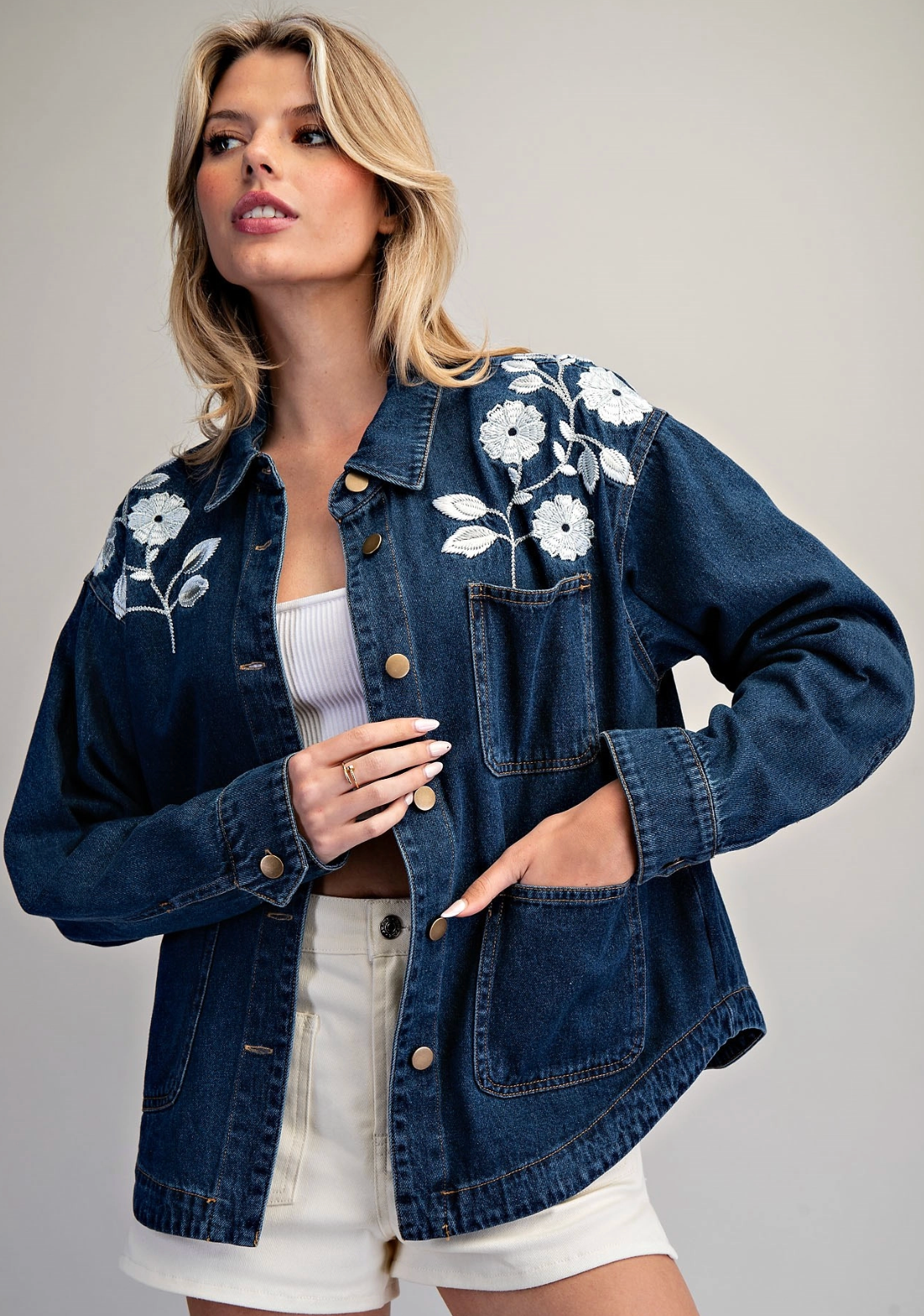 A woman wearing a dark blue denim jacket with white floral embroidery on the shoulders, styled over a white ribbed tank top and white shorts against a neutral gray background.
