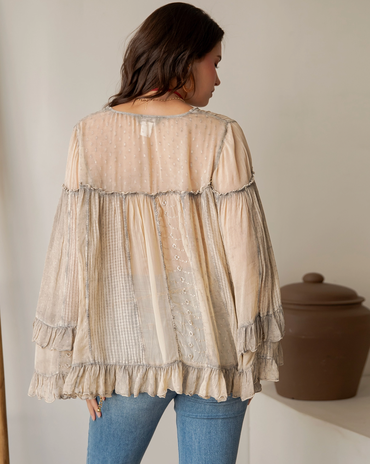 A woman shown from the back wearing a loose beige sheer blouse with layered ruffles and embroidered details, paired with blue jeans, in an indoor setting.
