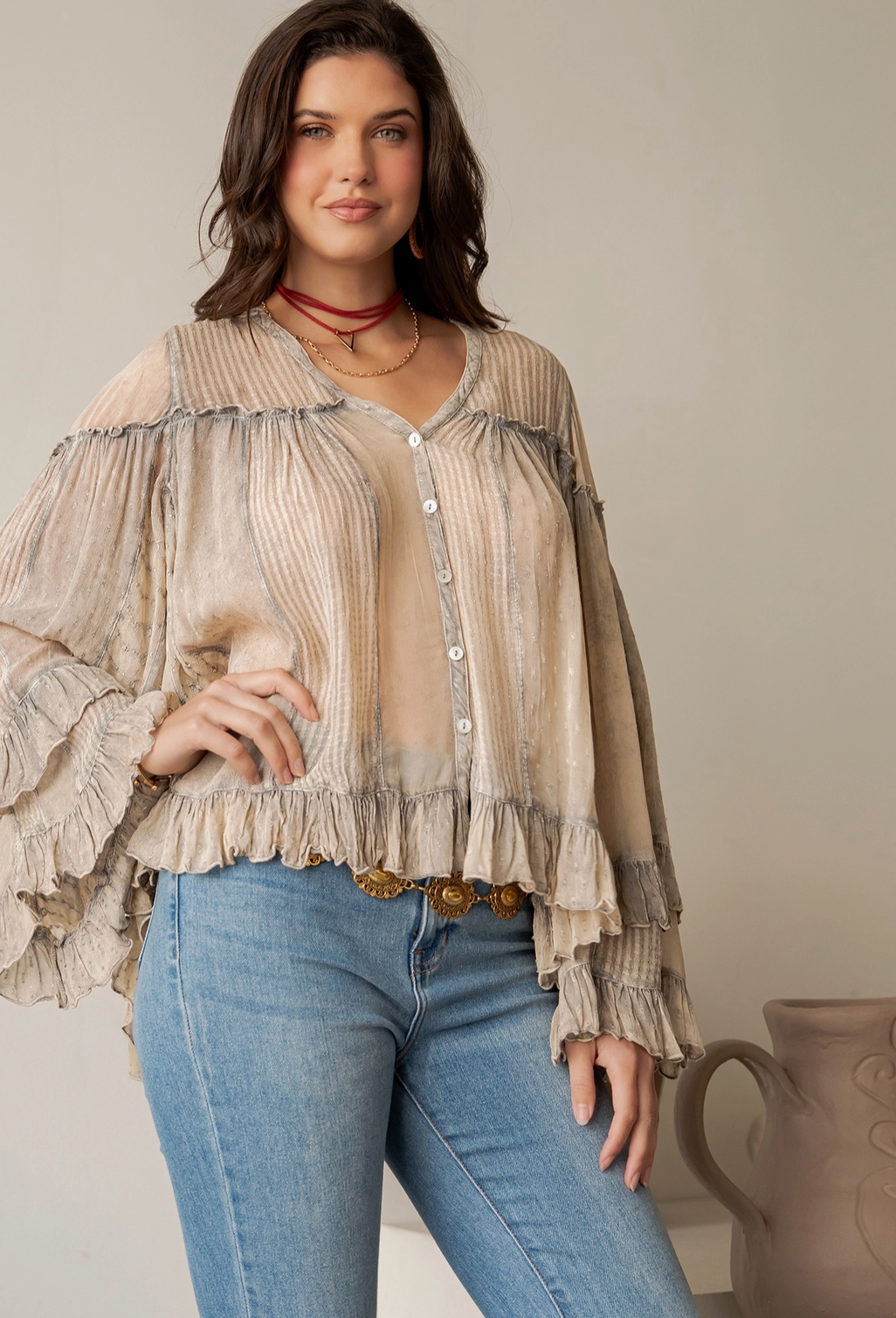 A woman models a beige and taupe ruffled button-up blouse with a sheer mesh insert, paired with blue jeans and gold-toned jewelry, set against a neutral studio background.