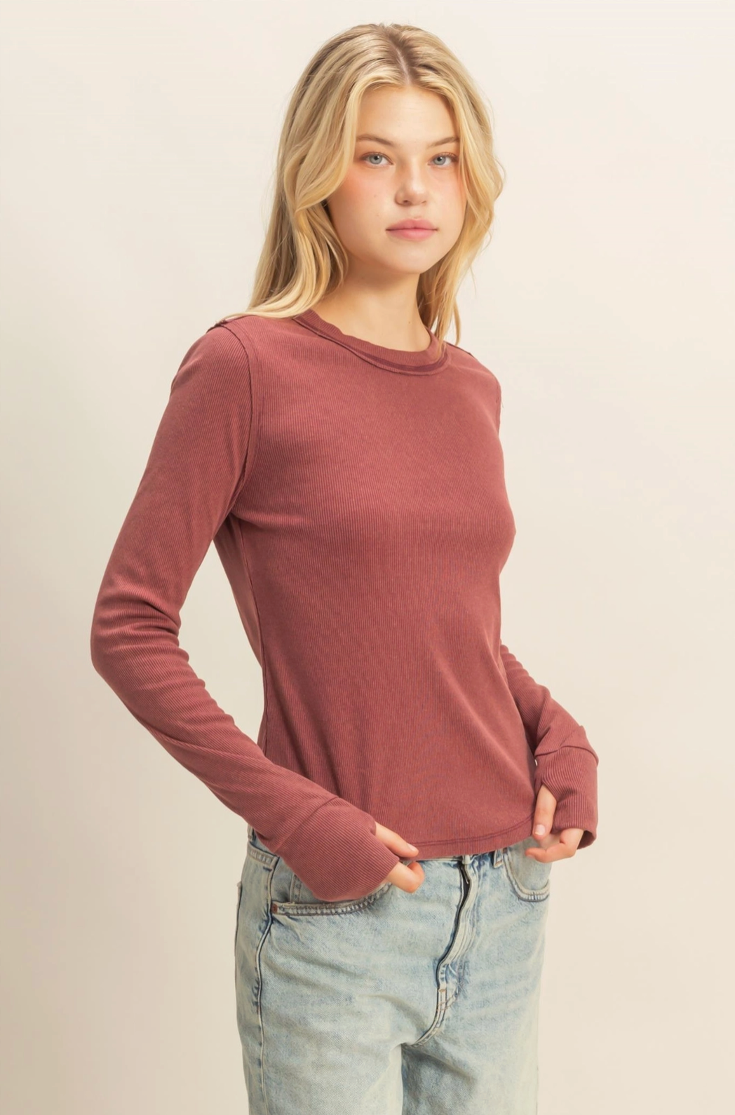 Woman wearing a maroon long-sleeve top and light blue jeans against a beige background