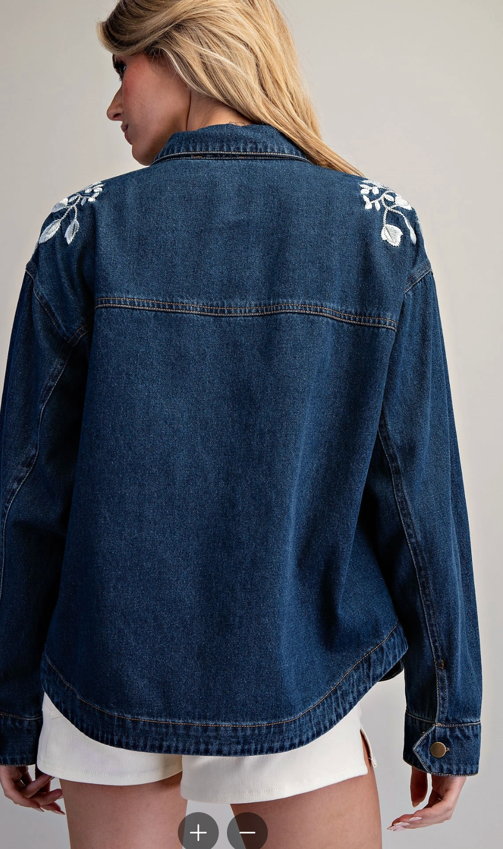 A woman viewed from the back wearing a dark blue denim jacket with white floral embroidery on the shoulders, paired with white shorts, against a light gray studio background.