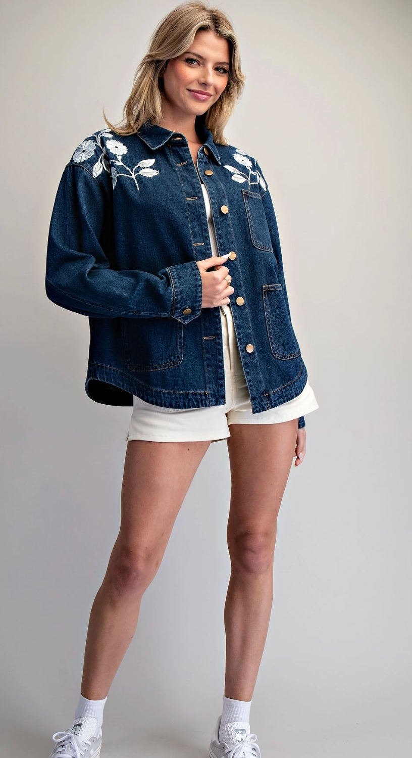 A woman modeling a dark blue denim jacket with white floral embroidery on the shoulders, paired with white shorts and white sneakers, against a light gray studio background.