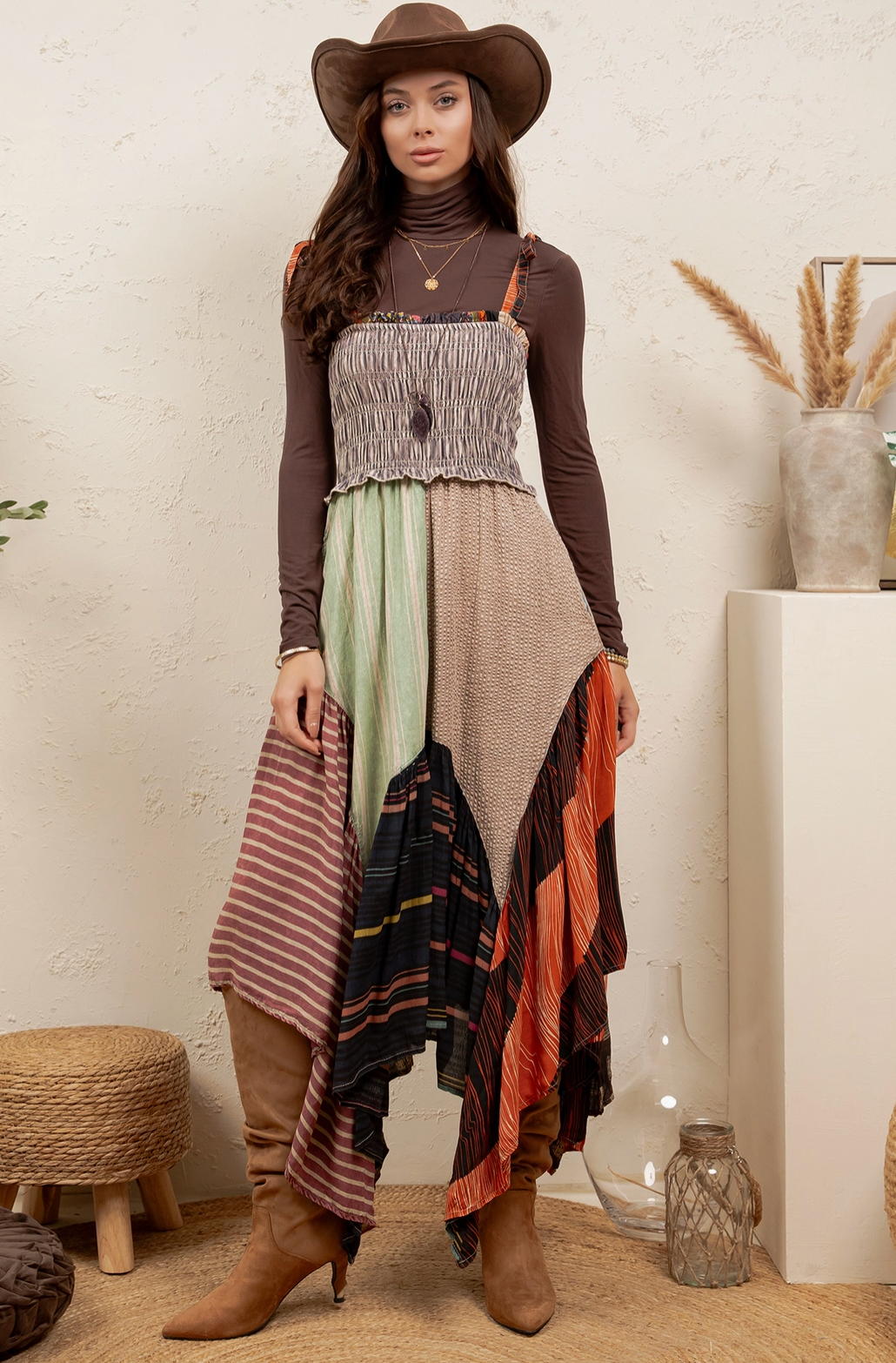 A woman wearing a bohemian-style patchwork maxi dress with a smocked bodice, long brown sleeves, and asymmetrical draped layers in beige, green, orange, and striped patterns. She is also wearing a brown wide-brim hat and brown knee-high boots, standing in a styled room with neutral-toned decor.
