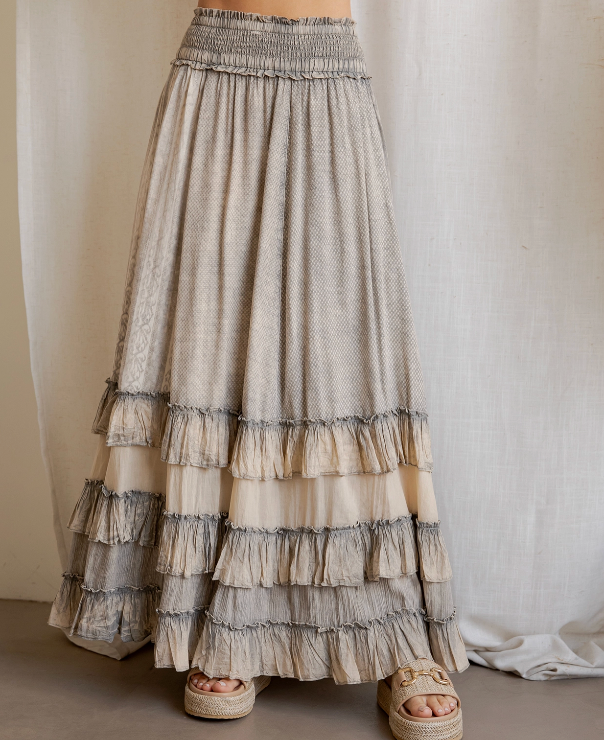 A long tiered maxi skirt in a textured beige and gray fabric, featuring an elastic waistband and multiple ruffled layers, shown on a model wearing woven espadrille sandals.