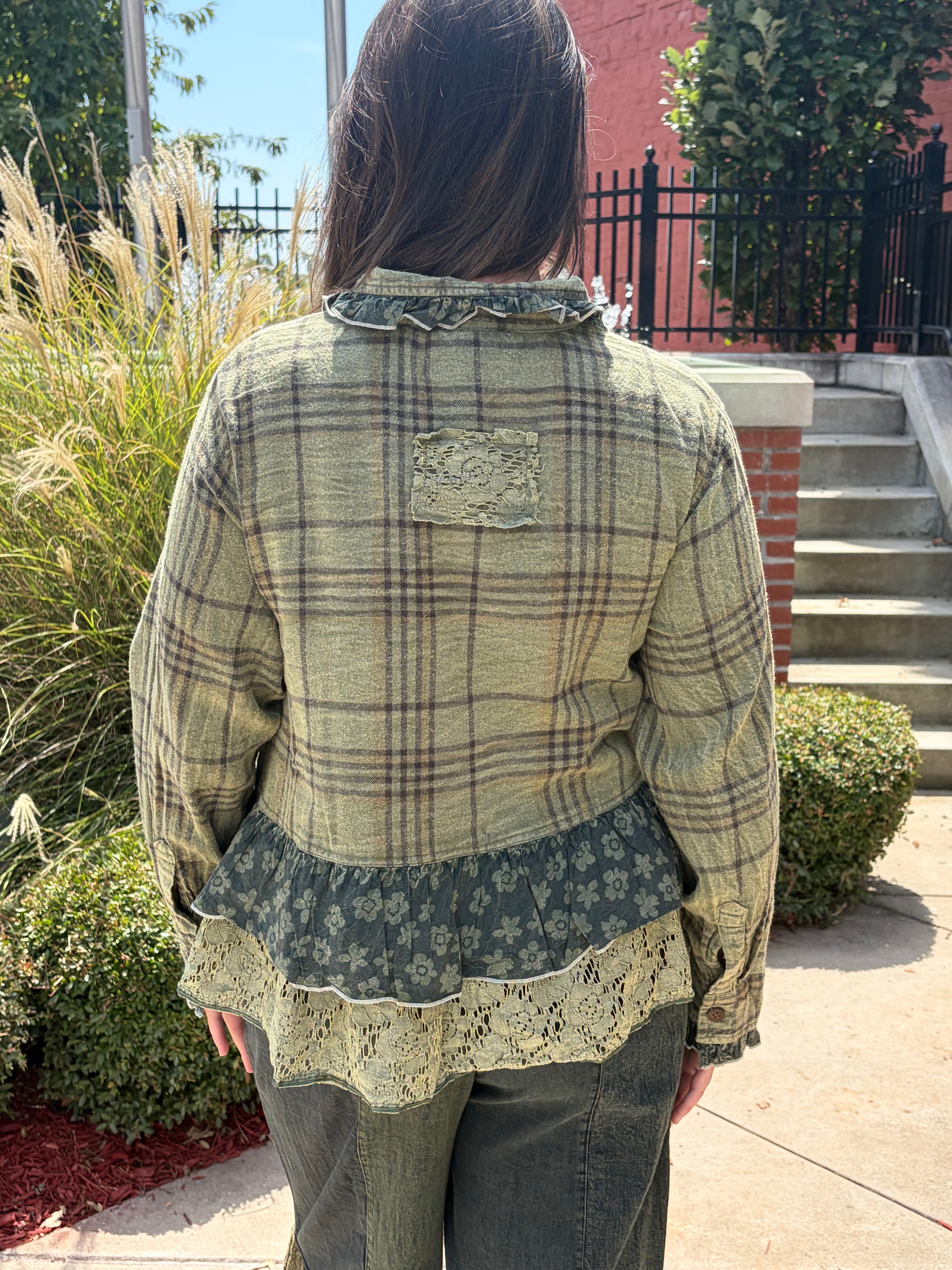 A woman viewed from behind wearing a vintage-style patchwork plaid shirt jacket in olive green, beige, and brown, standing outdoors on a sidewalk with plants and stairs in the background.
