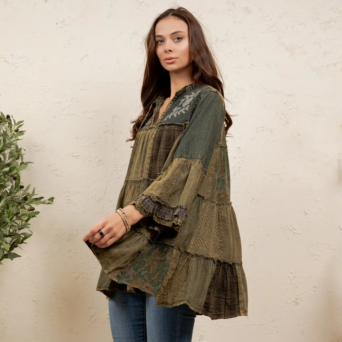 A woman wearing a bohemian-style tiered tunic top in olive green and brown patchwork fabrics with embroidery on the yoke, paired with blue jeans, standing against a textured beige wall.