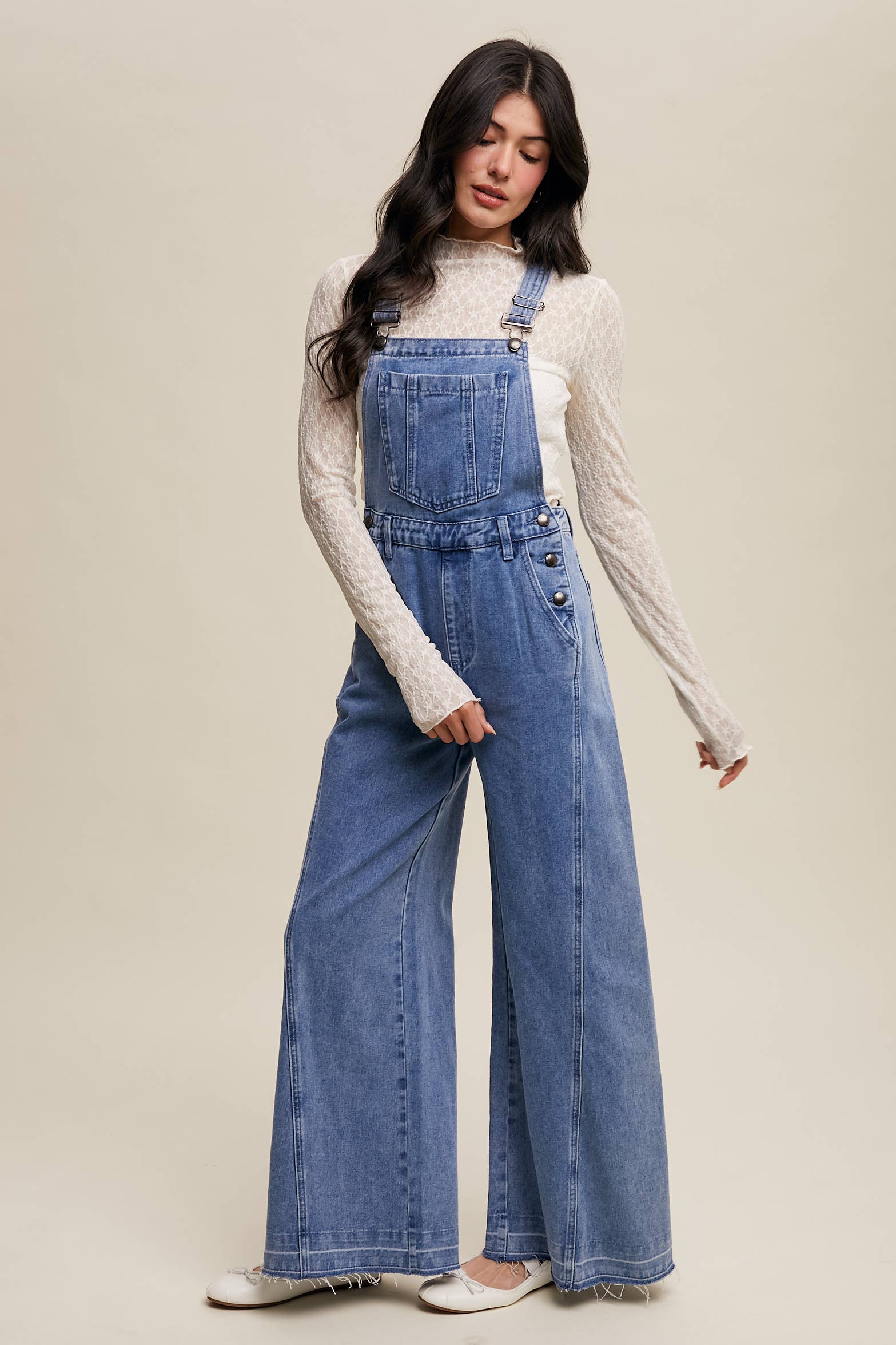 Light-wash wide-leg denim overalls with adjustable straps, front bib pocket, side buttons, and slant pockets, finished with a raw hem and layered over a cream long-sleeve top.
