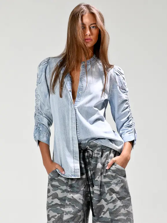 A woman modeling a light blue chambray button-up shirt with ruched sleeves and gray camouflage drawstring pants against a light gray studio background.