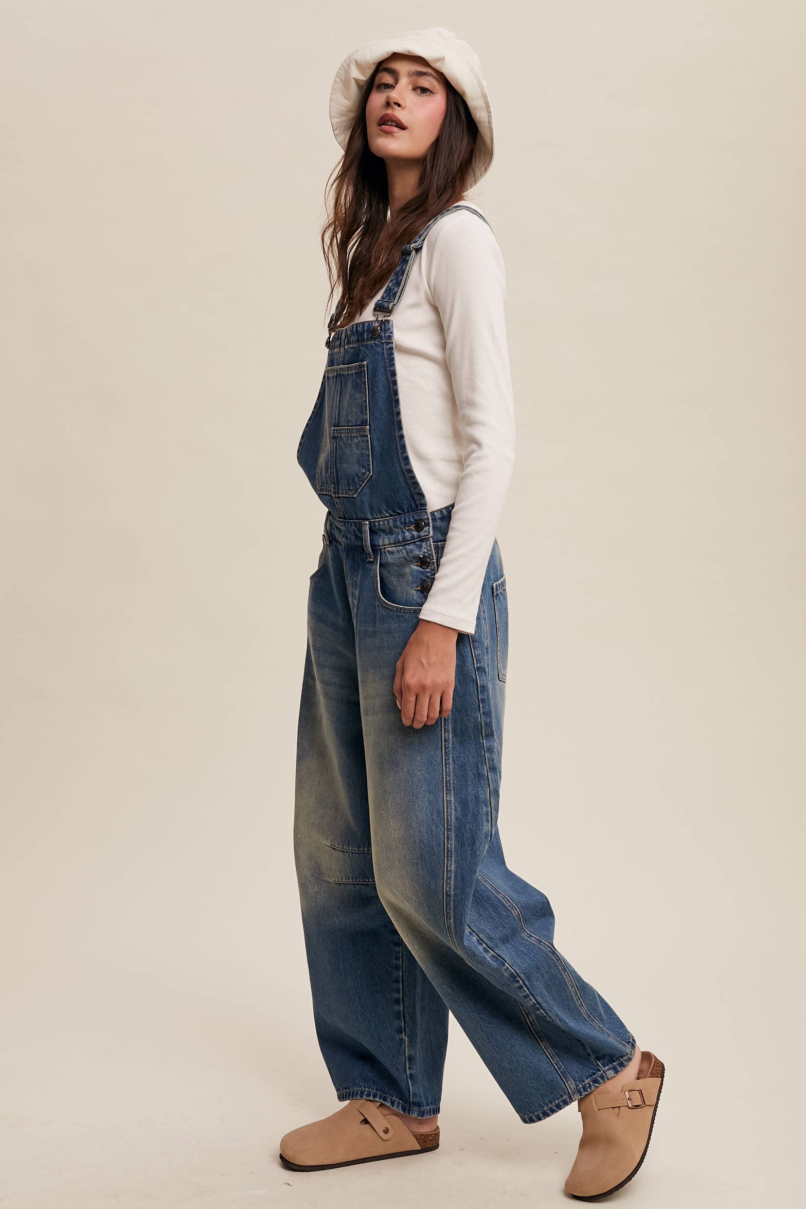 Side view- Vintage-inspired blue denim overalls with relaxed wide-leg fit, front chest pocket, and adjustable straps, styled with a white long-sleeve top and neutral clogs.
